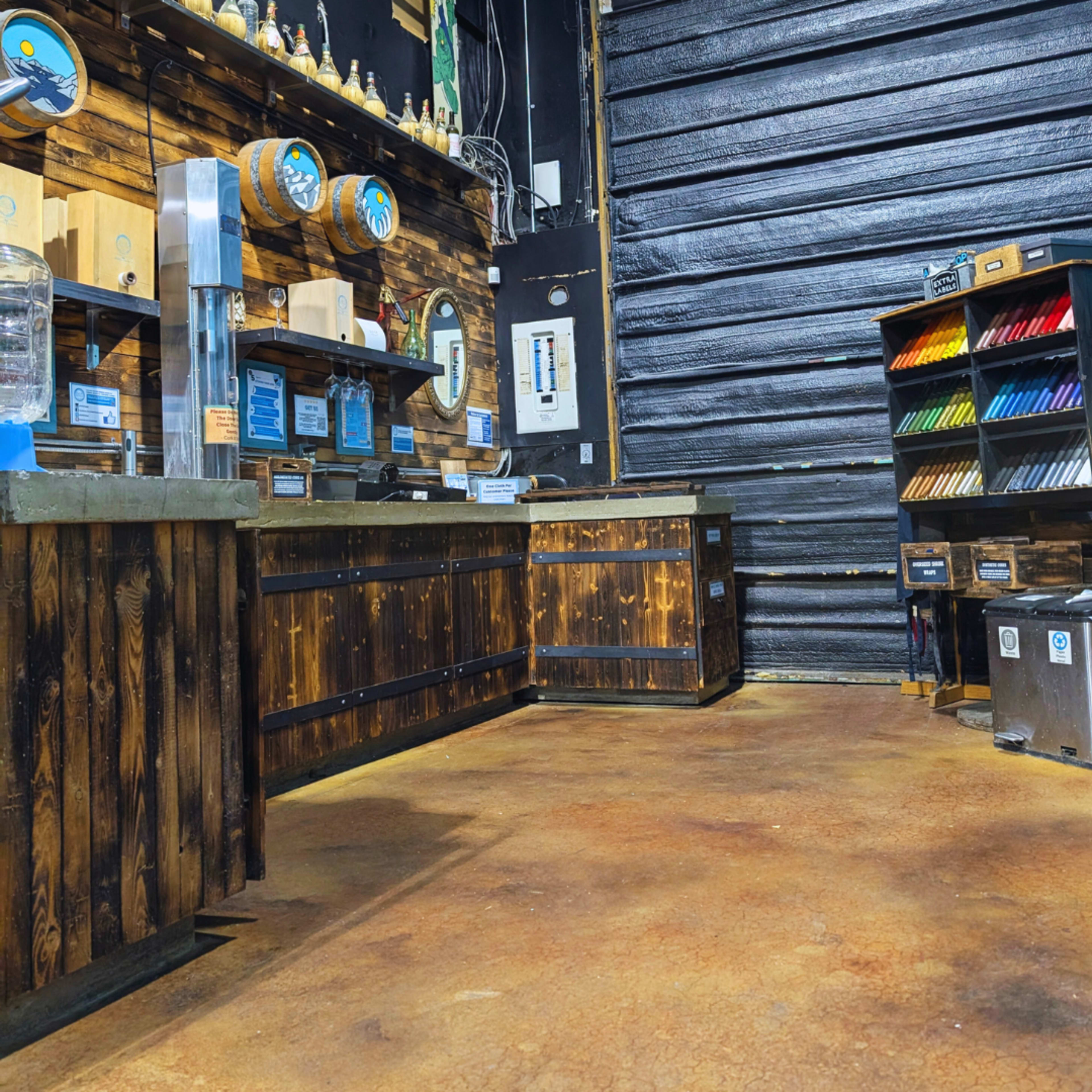 The interior of a store features a wooden counter with various displays and a wall of assorted colorful items beside a large metal door.