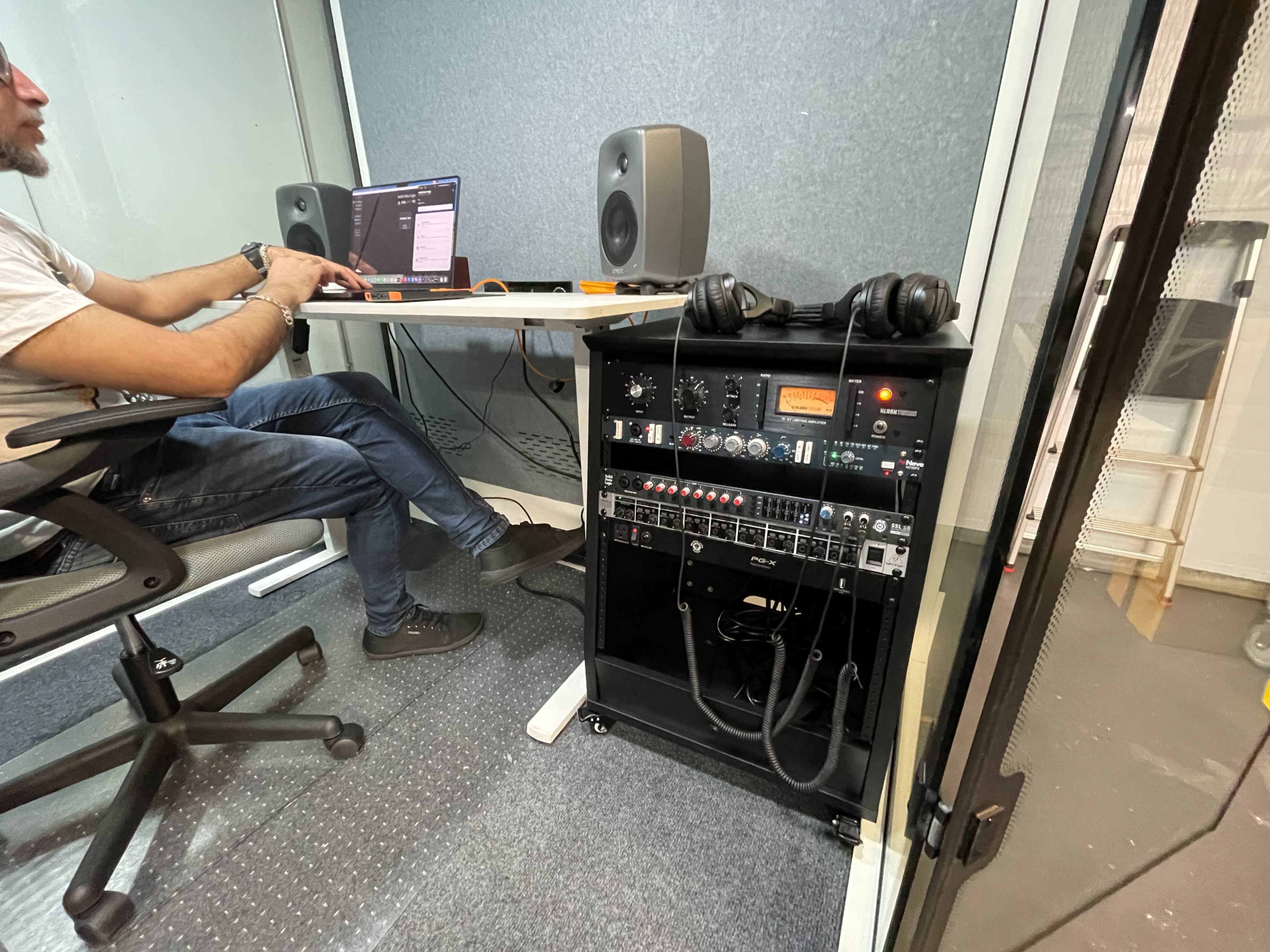 A person sits at a desk with a laptop and speakers in a soundproof room, while audio equipment is housed in a rack nearby.