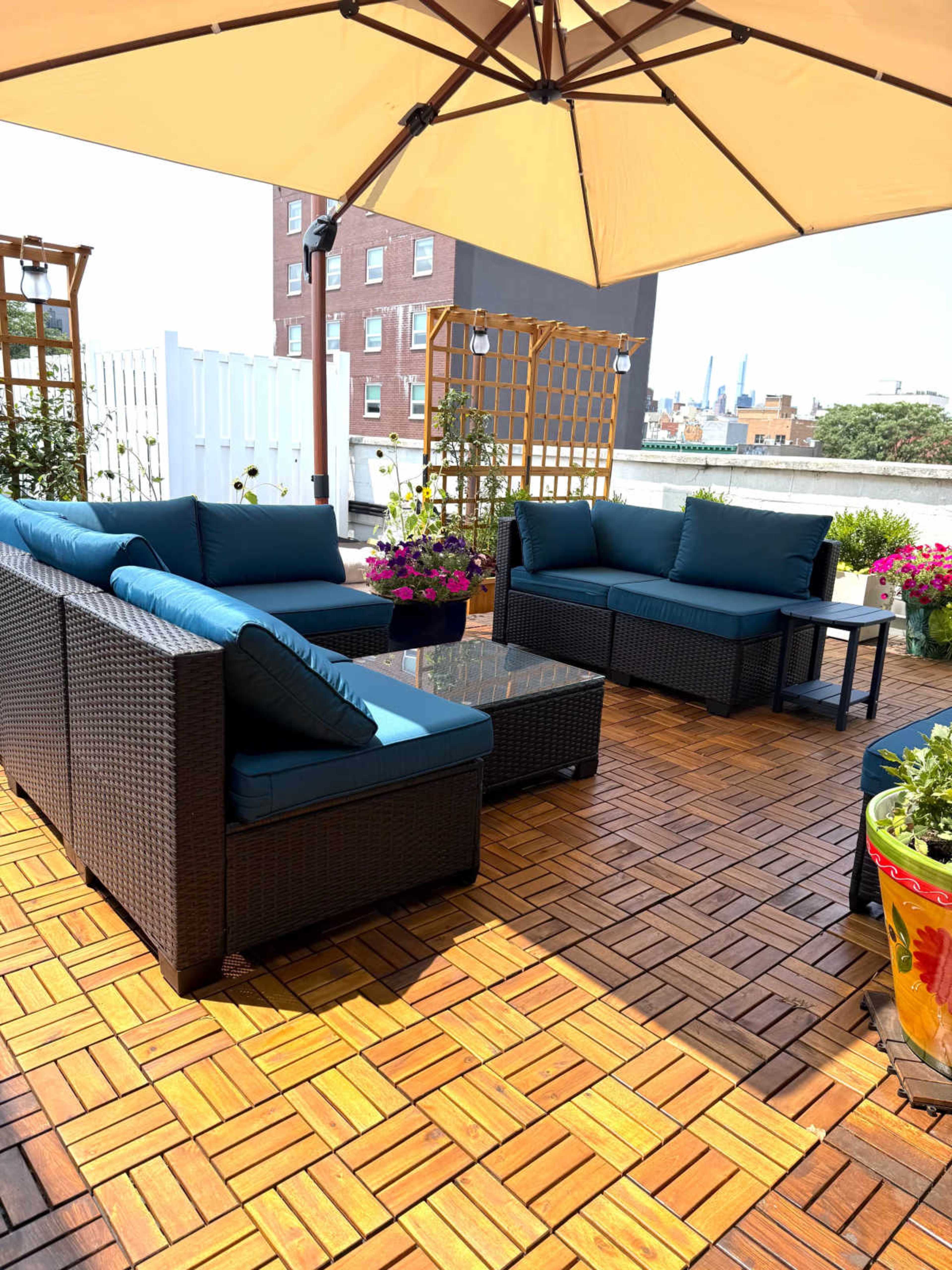 The image shows a rooftop terrace with wicker furniture, blue cushions, and colorful potted plants, all under a large umbrella.