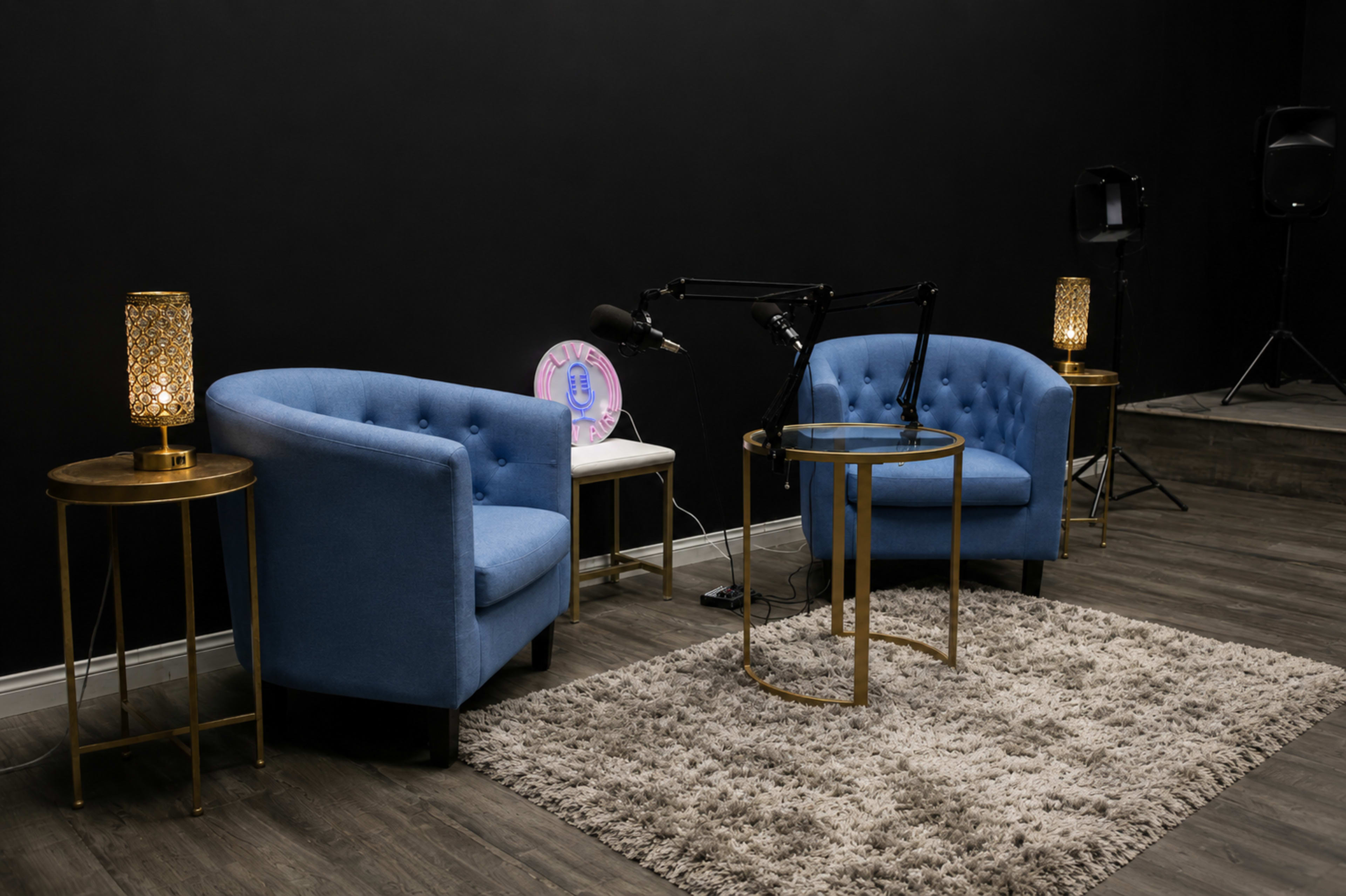 A modern interview setup features two blue upholstered chairs, a small round glass table, and decorative lamps on gold stands against a black wall.