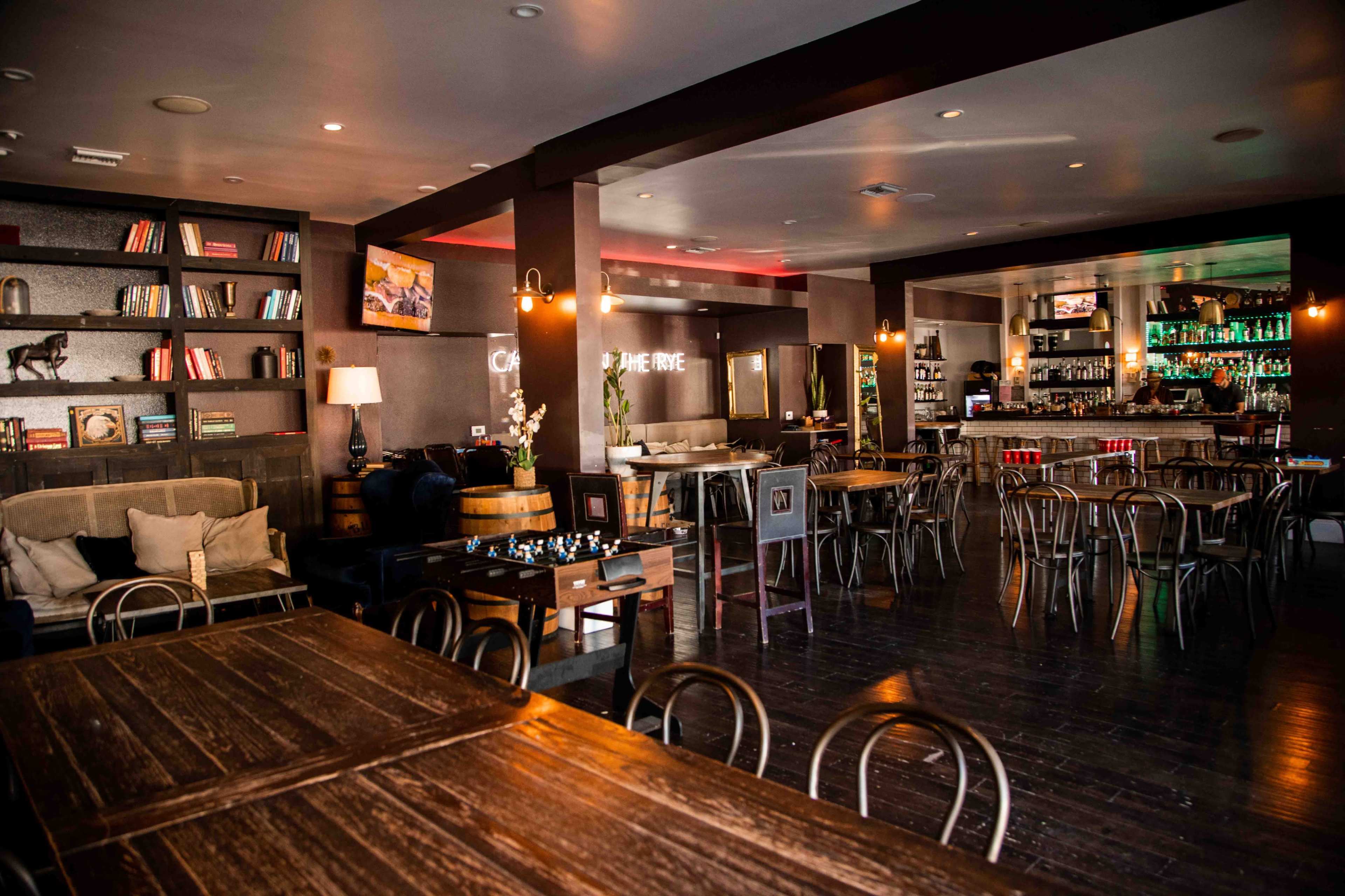 The image shows a spacious, modern bar and lounge area with wooden tables, a bookshelf, and a fully stocked bar in the background.