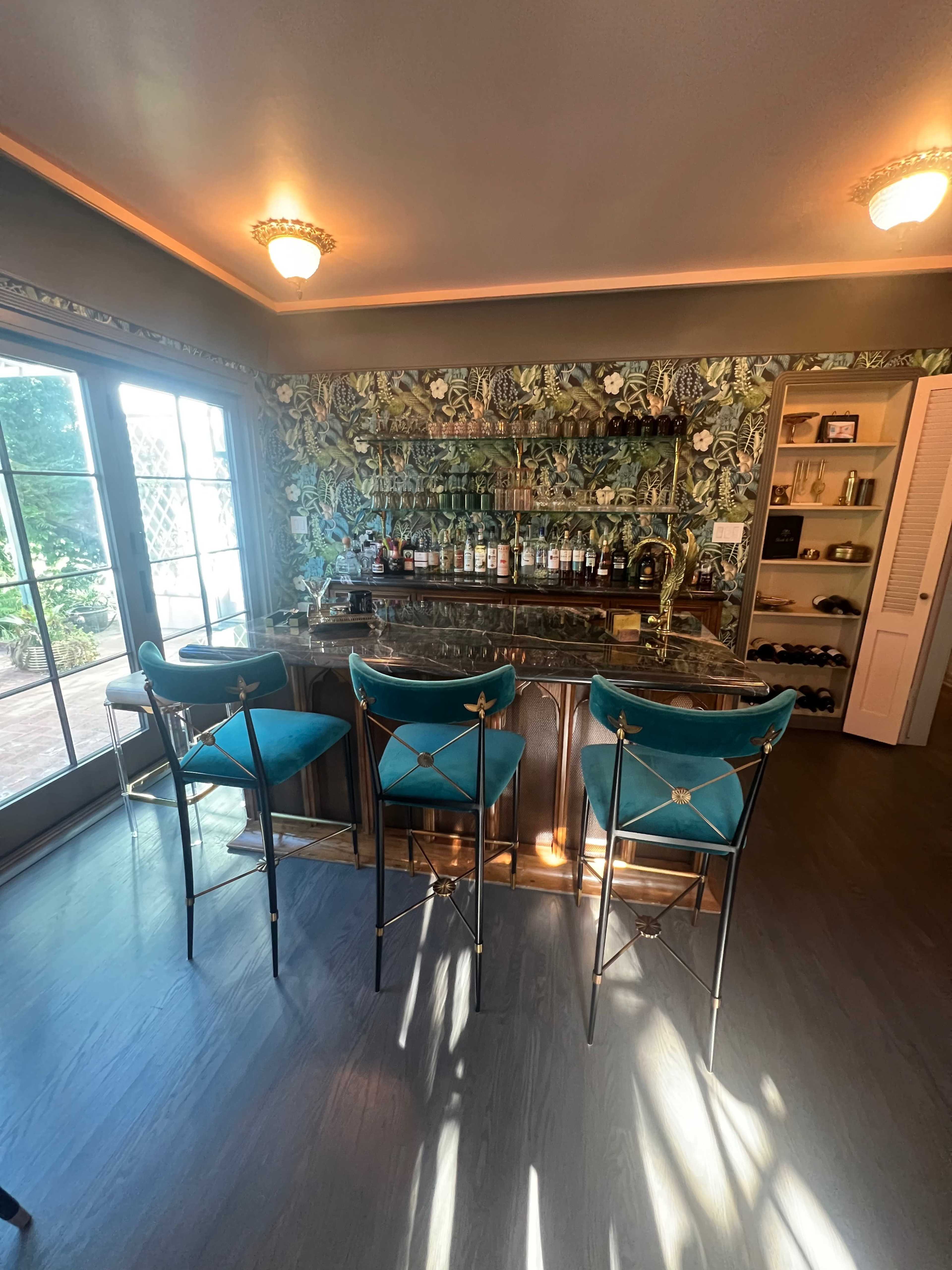 A home bar area with a marble countertop, four teal bar stools, and floral wallpaper behind a selection of bottles.