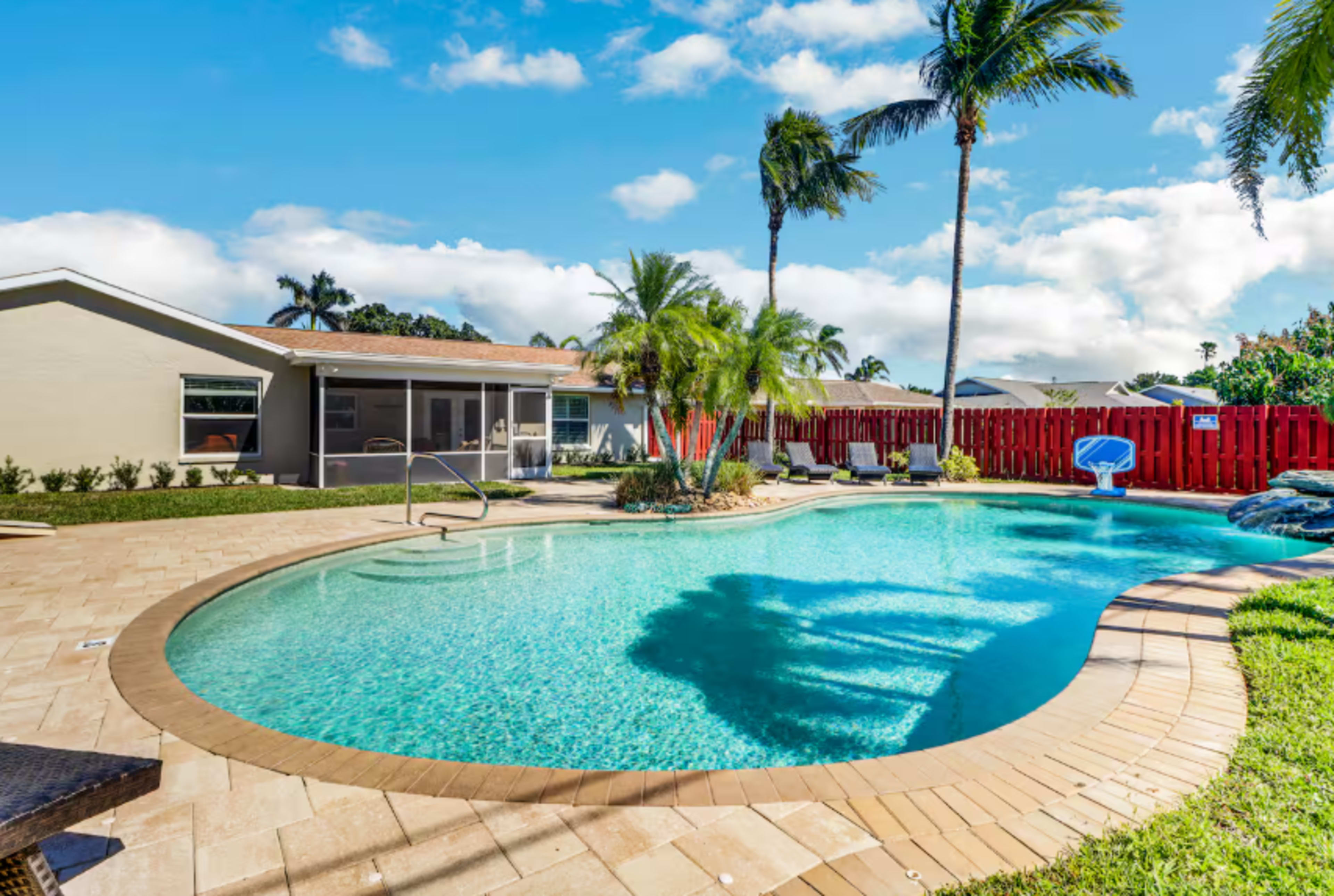 The image shows a residential backyard with a swimming pool surrounded by palm trees and a red wooden fence.