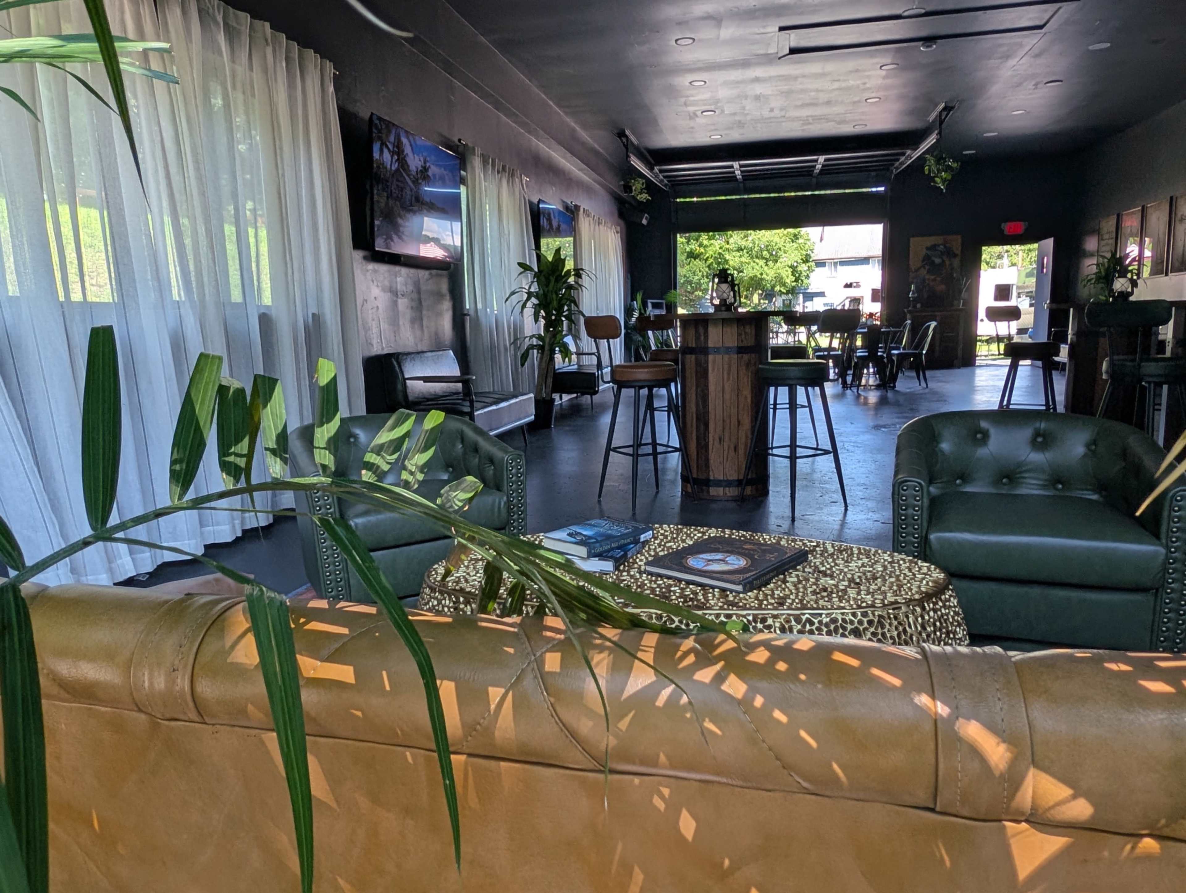 The image shows a modern, stylish interior of a lounge with large windows, green plants, leather and upholstered seating, and a bar area.