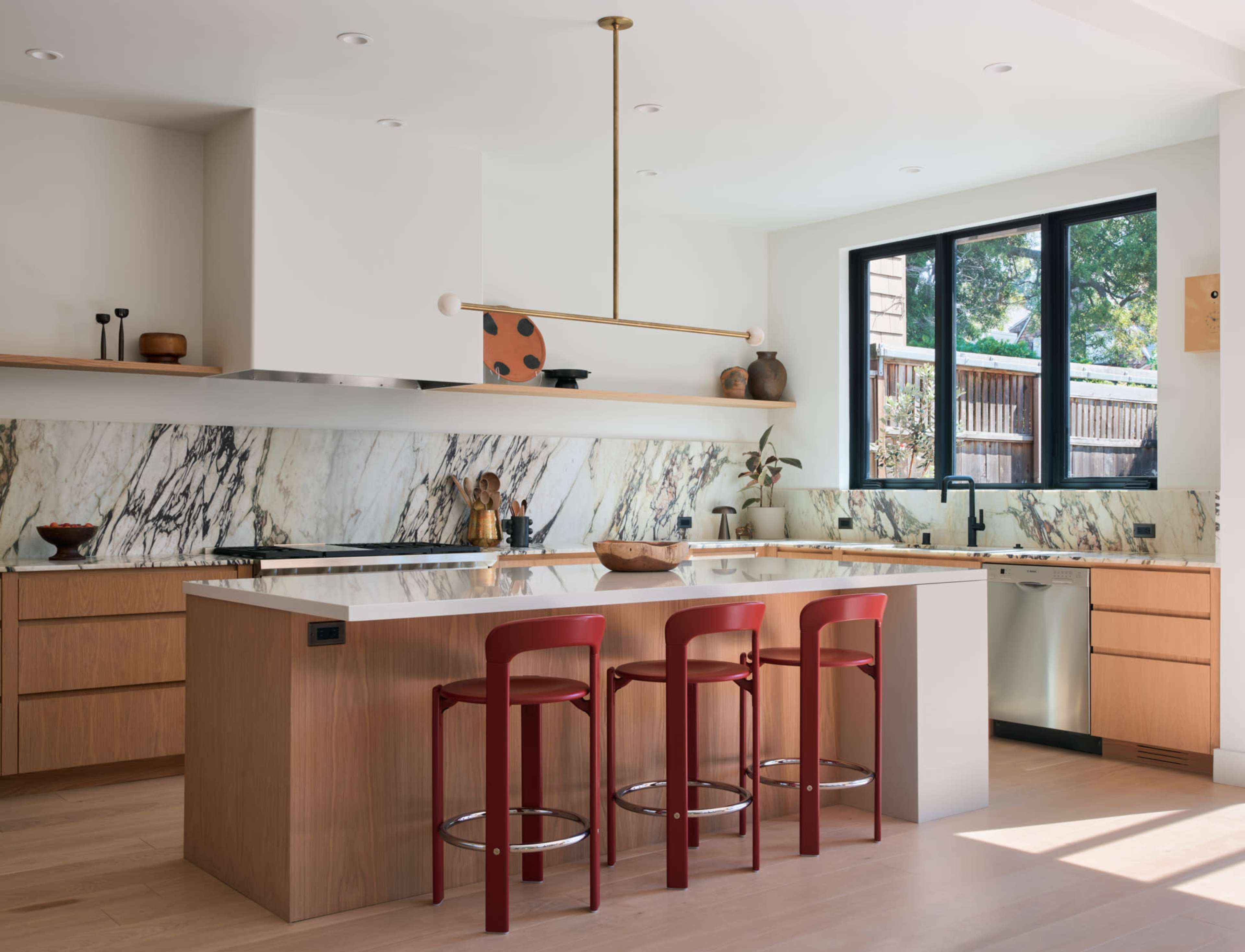 The modern kitchen features marble countertops, wooden cabinetry, and red bar stools, with large windows providing natural light.