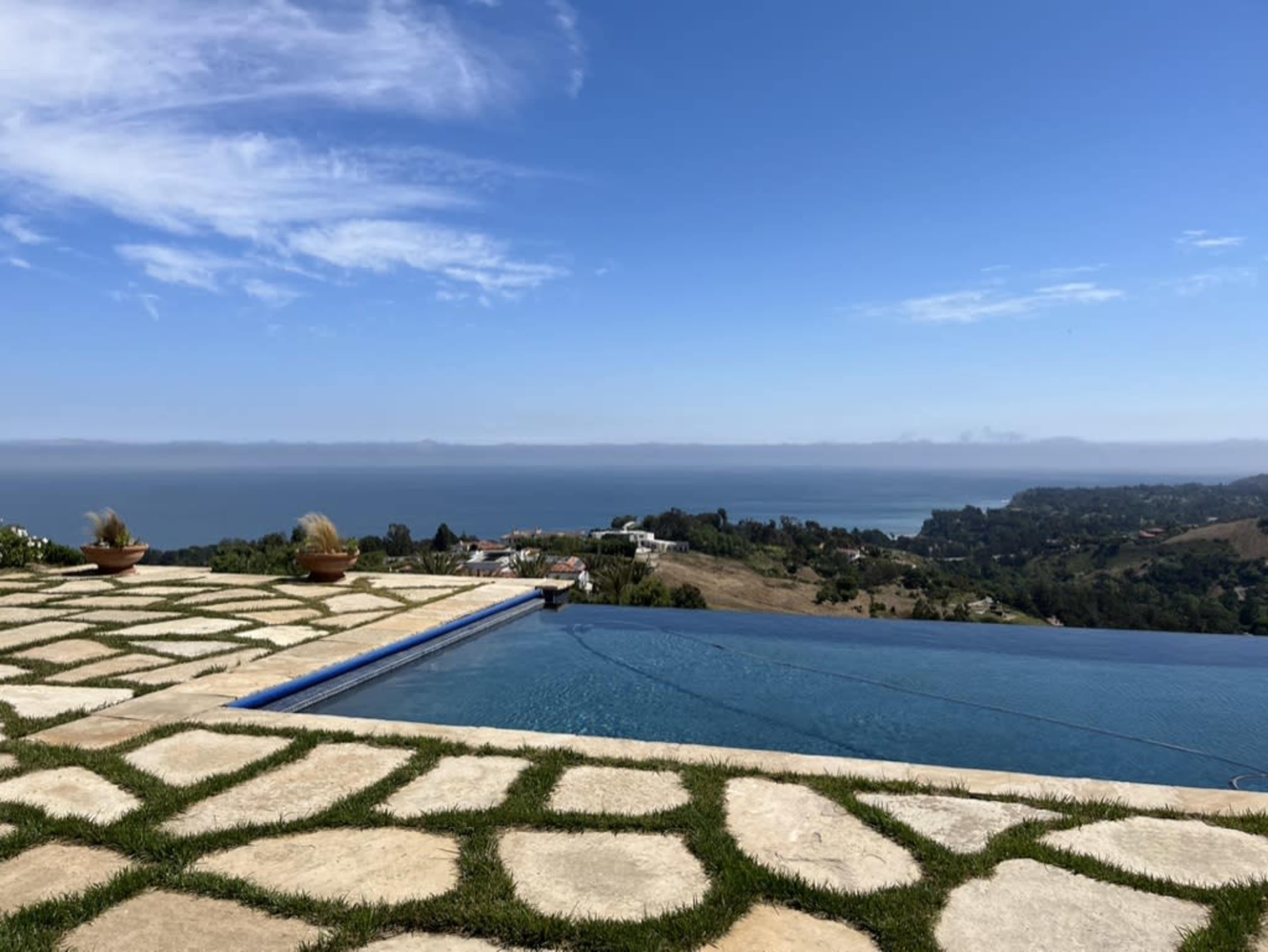 A swimming pool with an infinity edge overlooking a coastal landscape and the ocean under a clear blue sky.