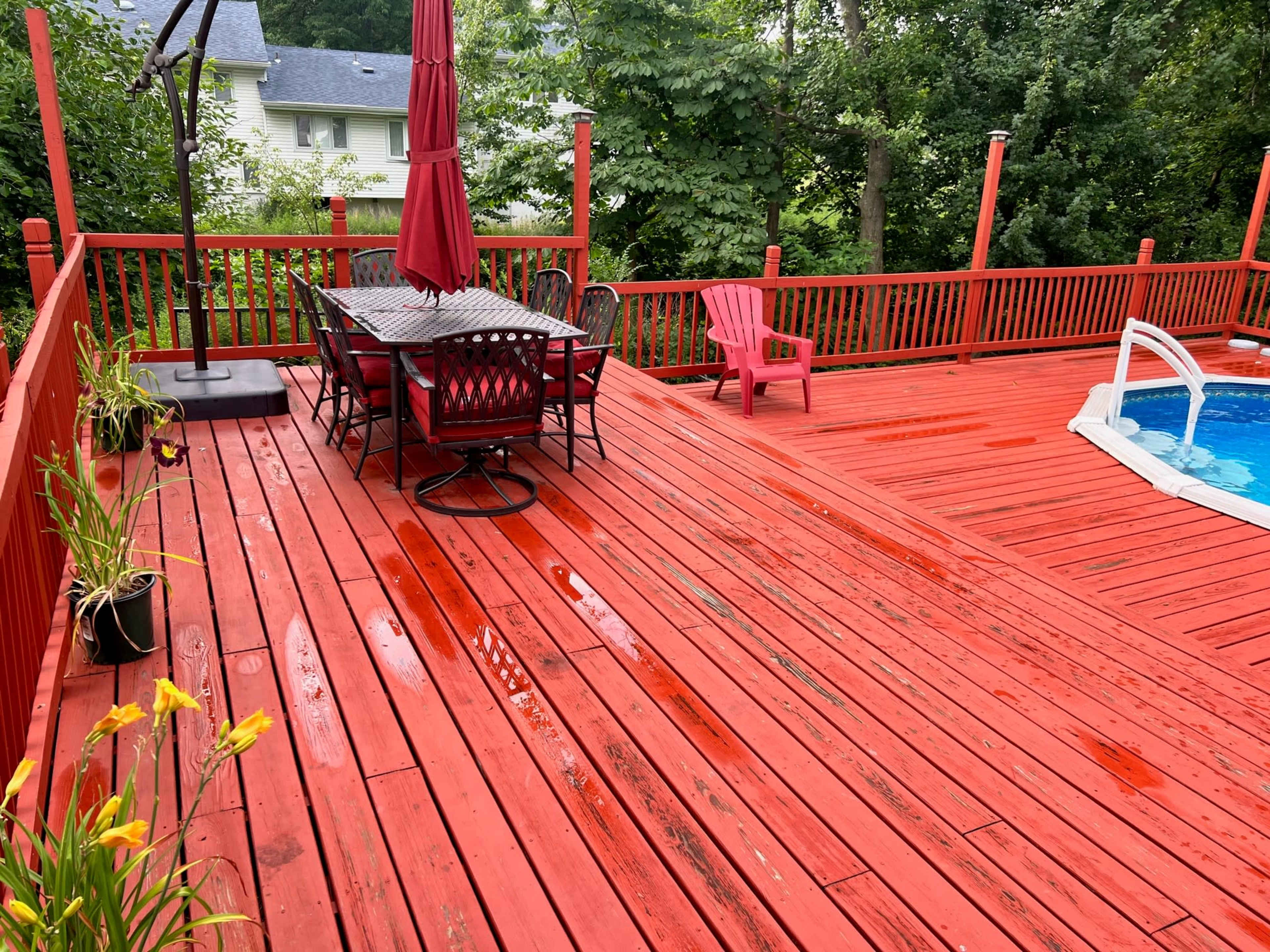 A red wooden deck features a dining table with chairs, an umbrella, and a pool, surrounded by green trees.