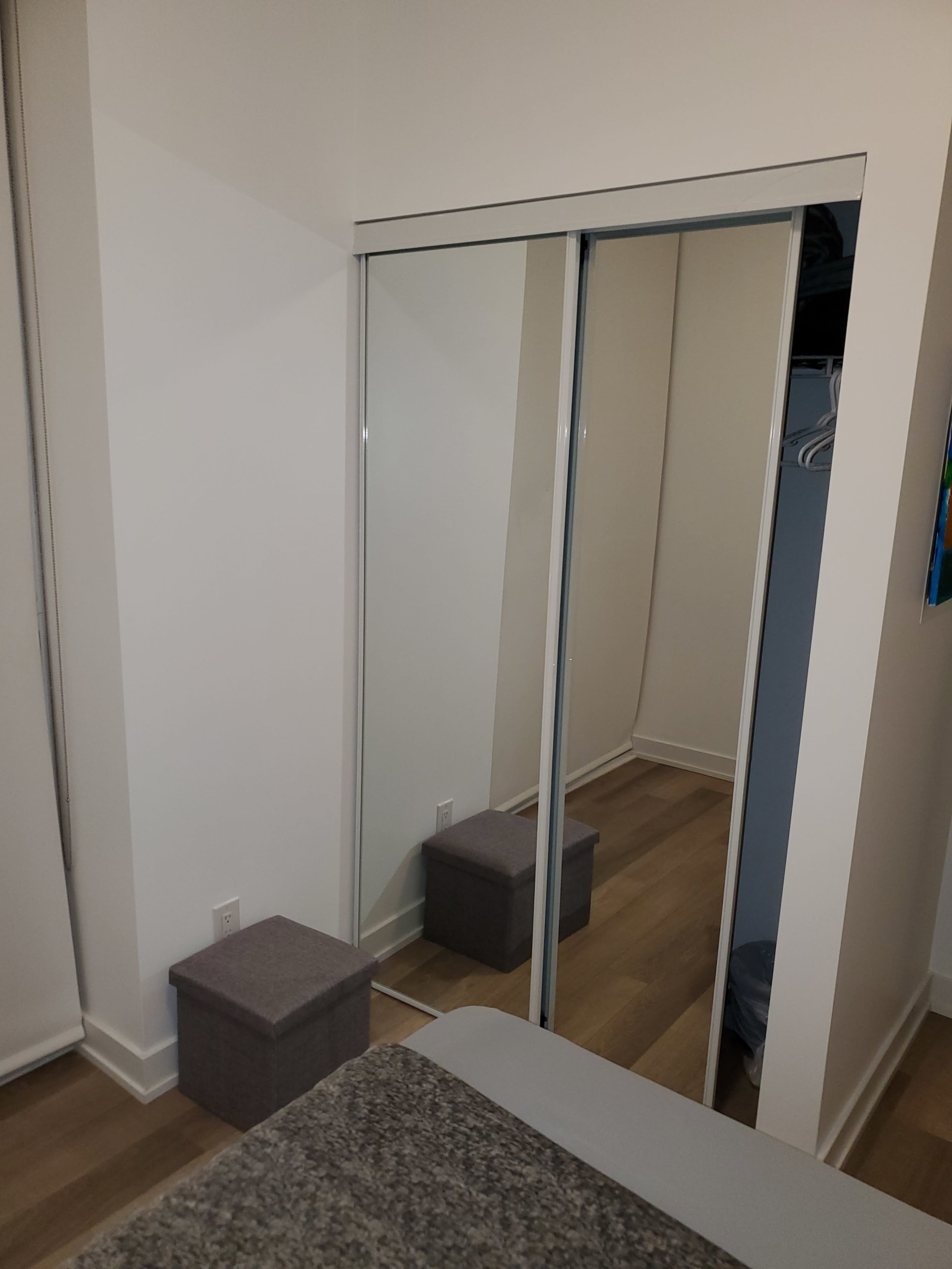 A bedroom with a bed in the foreground, a gray storage box near the foot of the bed, and a mirrored closet door against a light-colored wall.