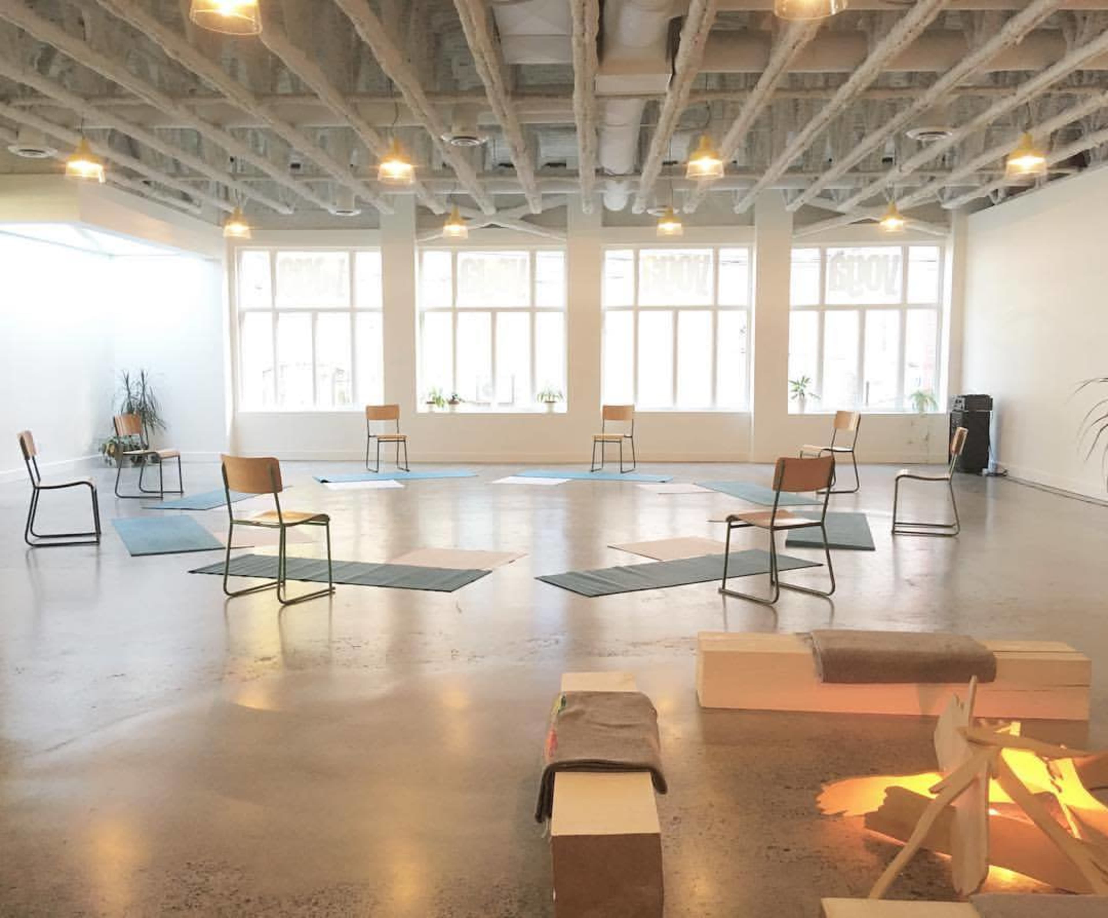 A spacious, well-lit room with several chairs arranged in a circle on a concrete floor, accompanied by mats and a small seating area.