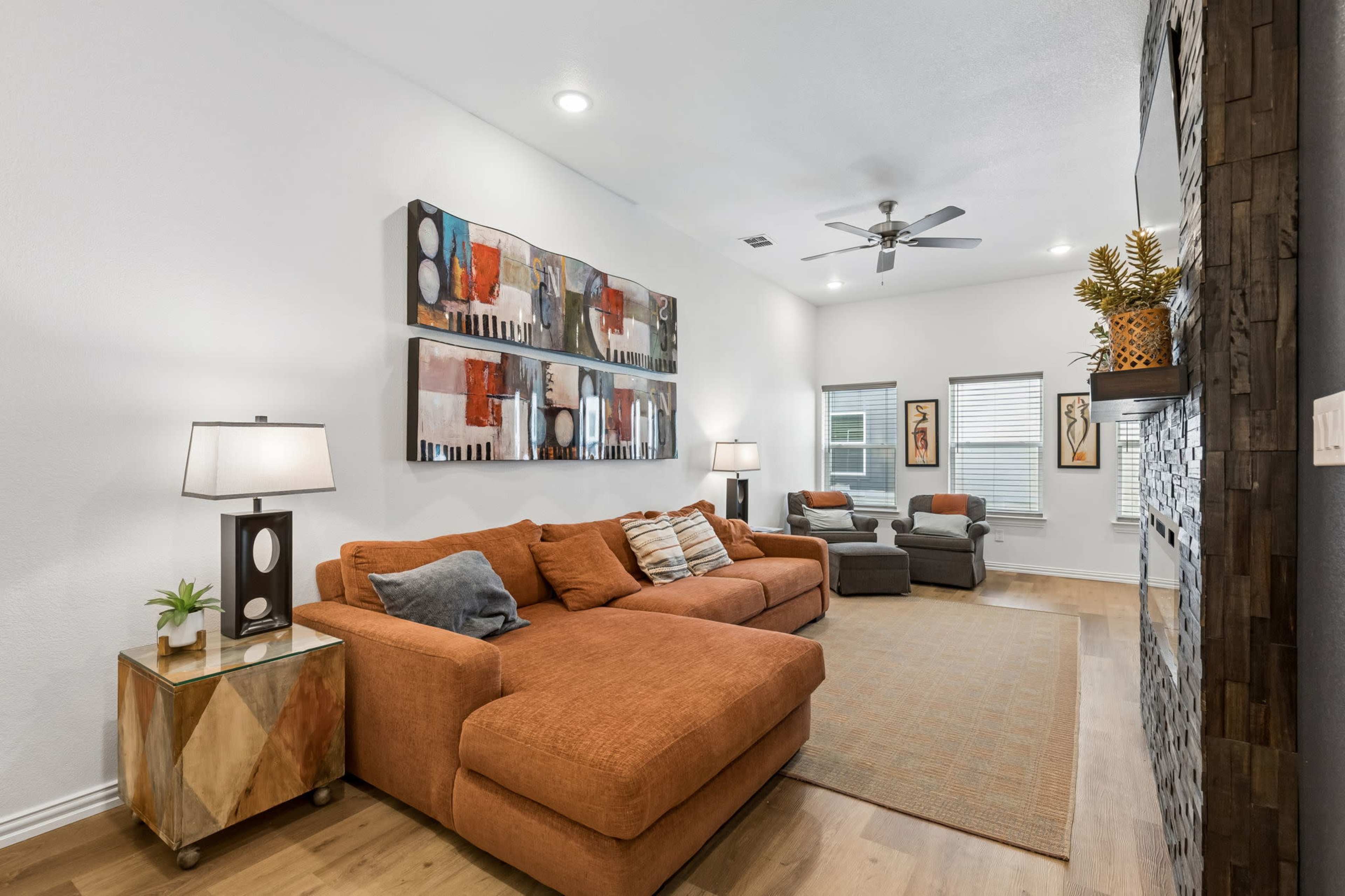 The living room features an orange sectional sofa, a wooden coffee table, abstract wall art, and a stone accent wall, with windows allowing natural light.