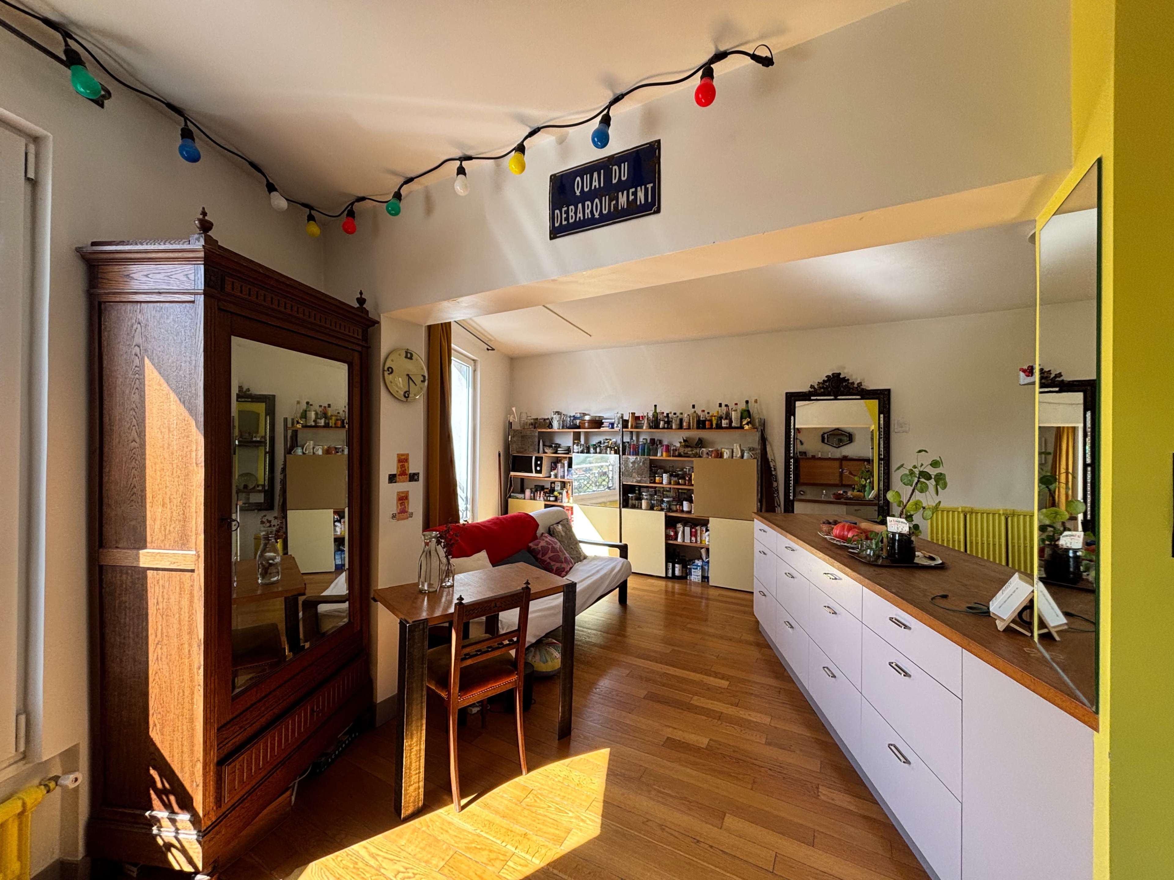 The image shows a bright, open living space with a wooden floor, a large mirror, a dining table, and a kitchen area featuring white cabinetry and a shelf filled with bottles.