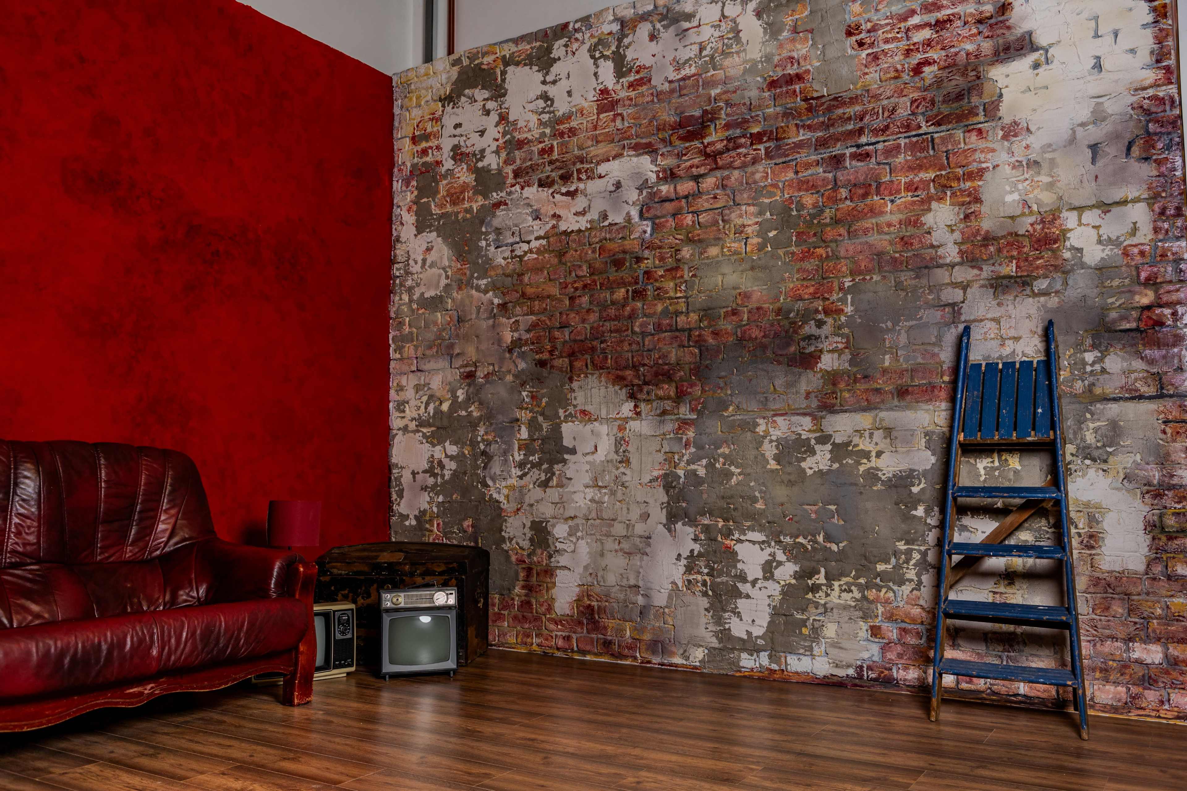 The image shows a corner of a room with a red wall, a weathered brick wall, a brown leather couch, a vintage television, and a blue wooden ladder.