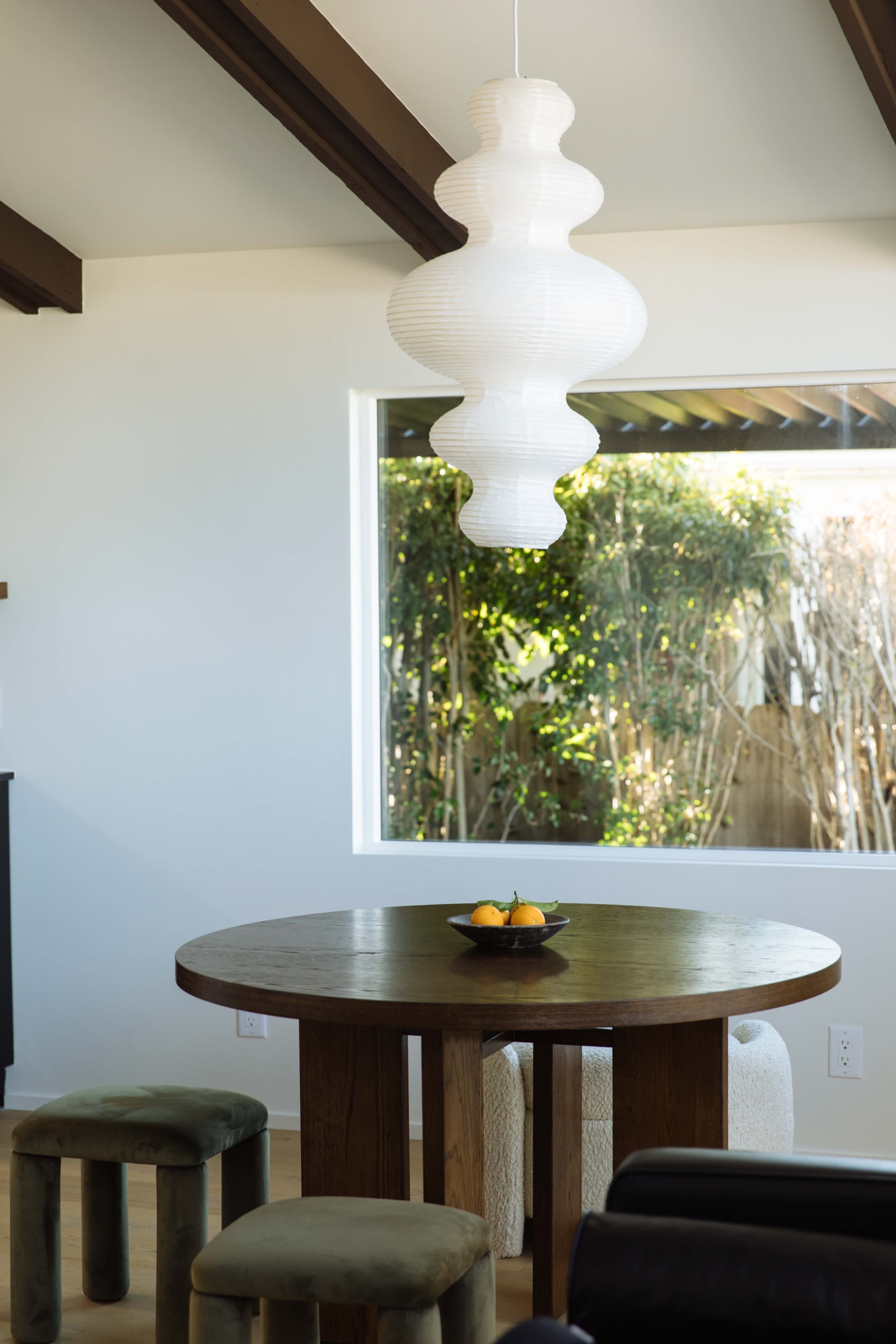 A round wooden table with a bowl of oranges sits beneath a white pendant light in a well-lit dining area with a large window.