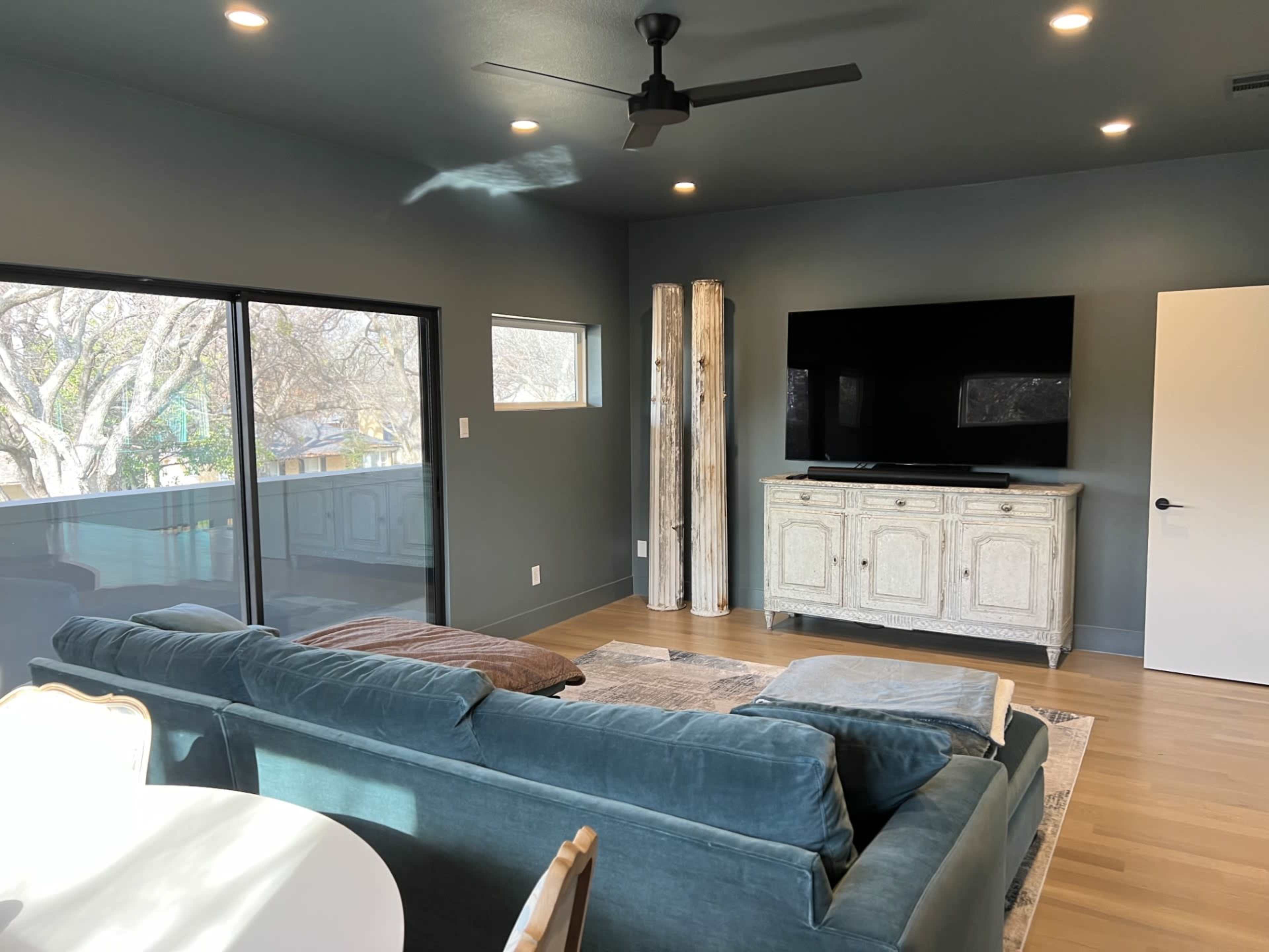 A living room features a blue sofa, a wooden TV stand, and large windows that provide a view of the outdoors.