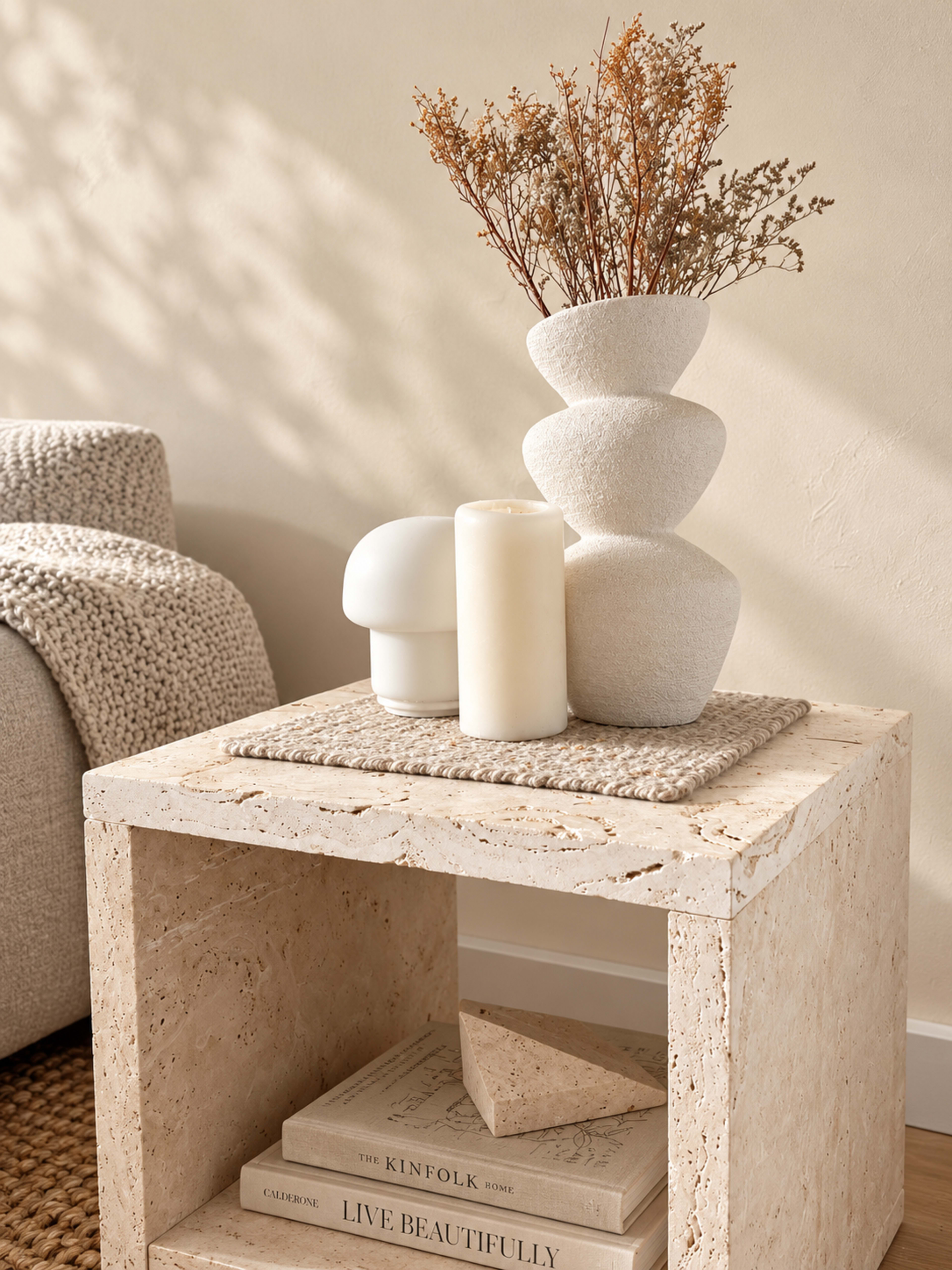 The image shows a textured side table made of stone with a stacked vase, two candles, and a small decorative object on top, accompanied by artfully arranged dried flowers.