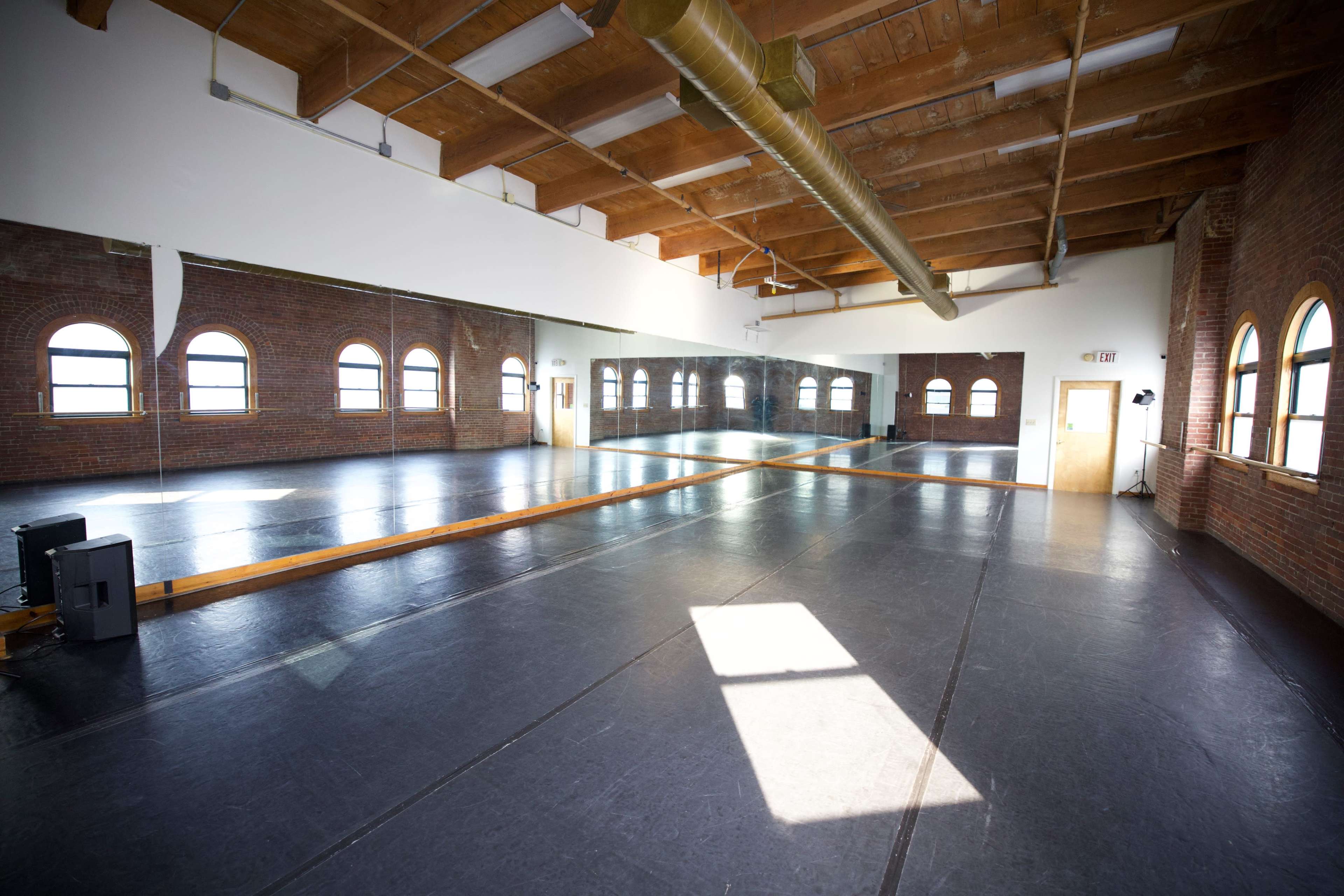 A spacious dance studio with wooden beams, large windows, and a mirrored wall.