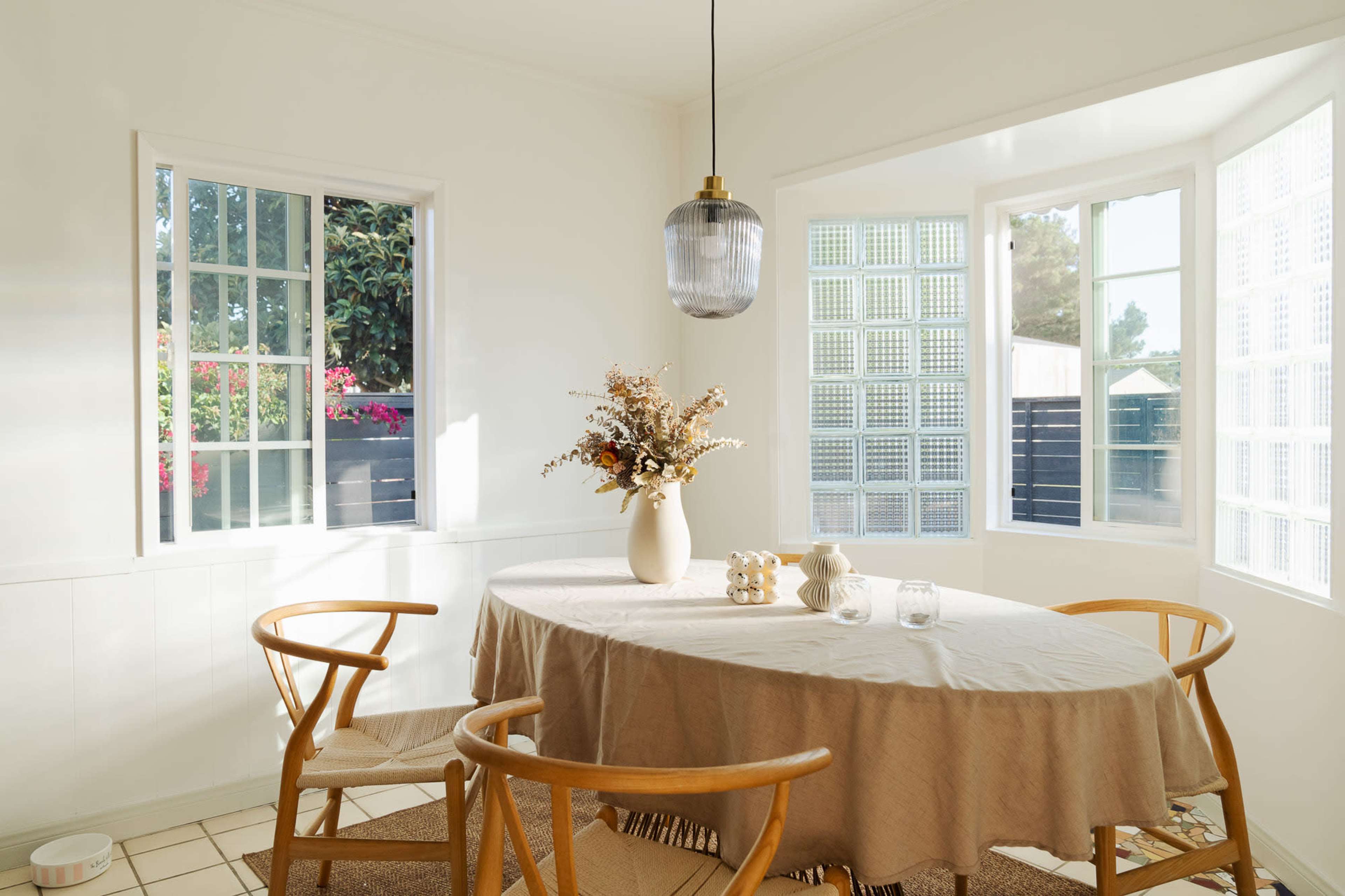 A well-lit dining area features a round table with a beige tablecloth, surrounded by wooden chairs, and a vase of dried flowers at the center.