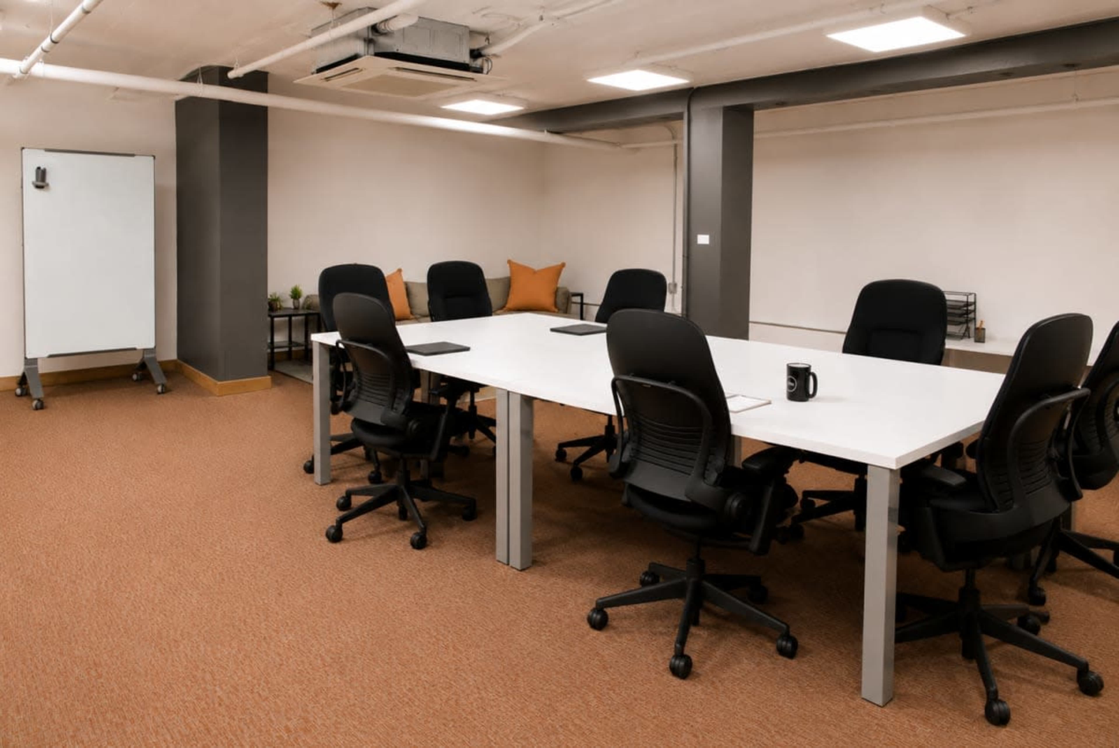 The image shows a modern conference room with a long white table surrounded by black chairs, a whiteboard on one side, and decorative pillows on the floor.