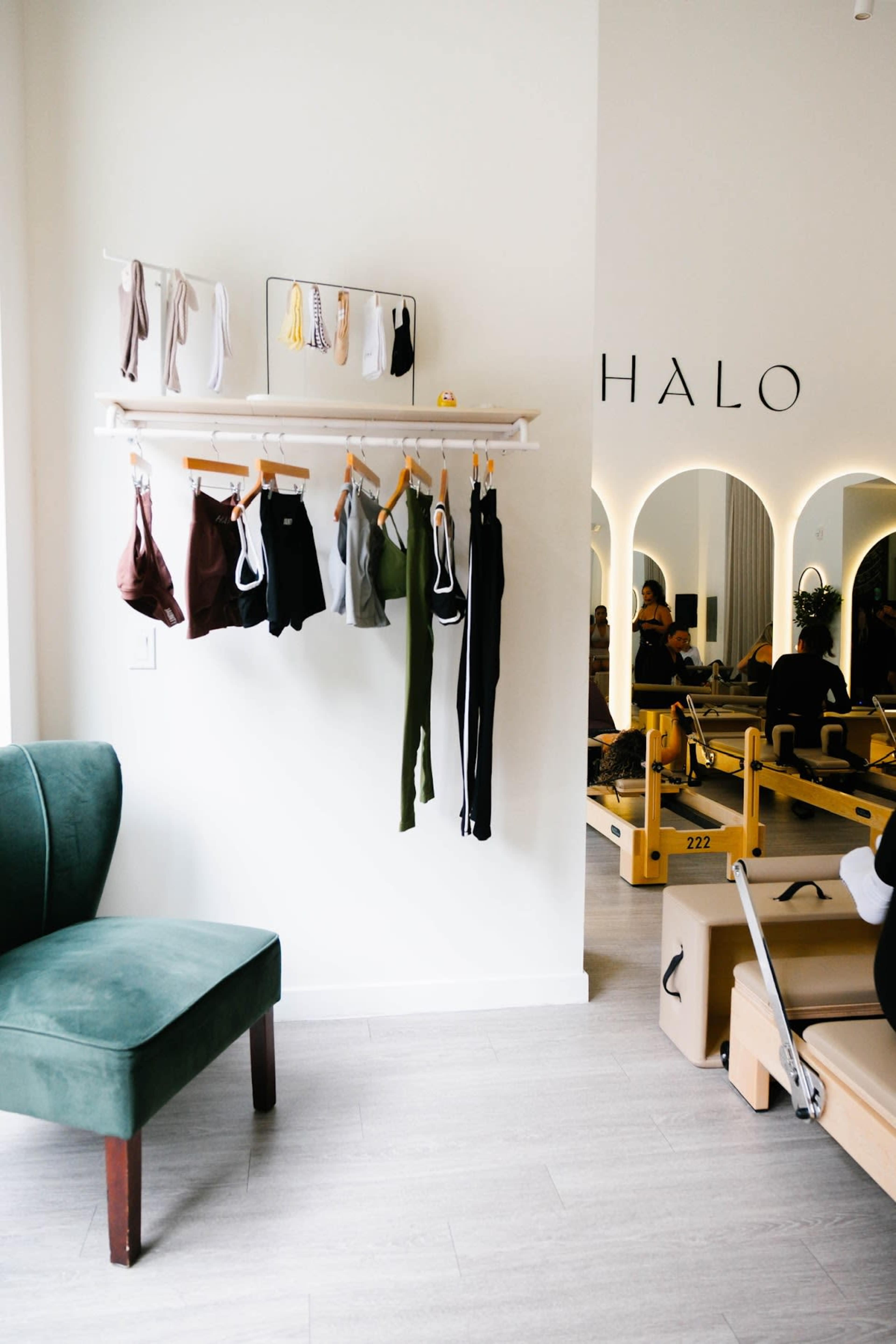 The image shows a fitness apparel display on a wall with various styles of clothing hanging from wooden hangers, alongside a green chair and a fitness studio in the background.