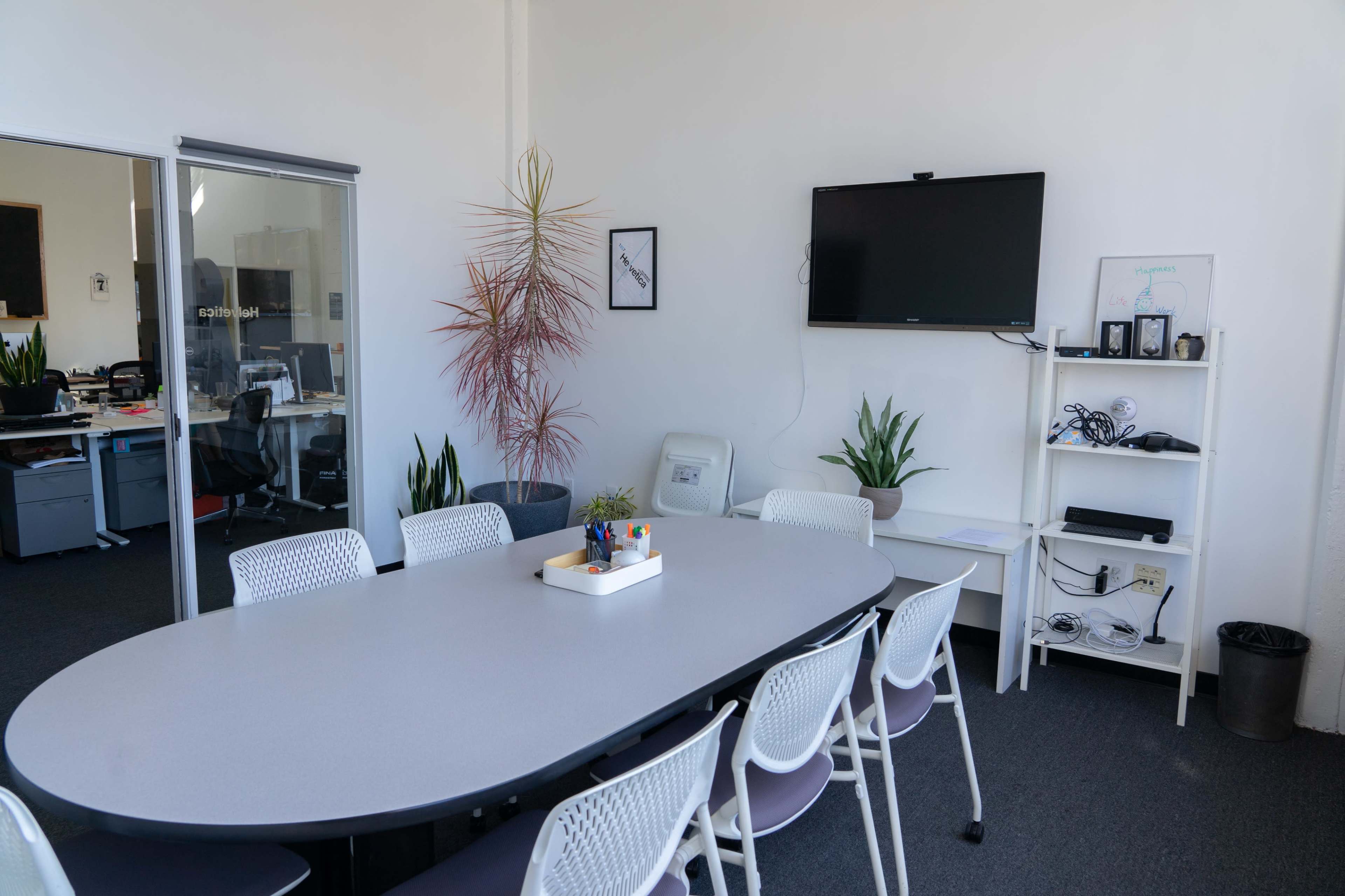 The image shows a modern meeting room with a long oval table, several chairs, a television mounted on the wall, and plants in the corners.