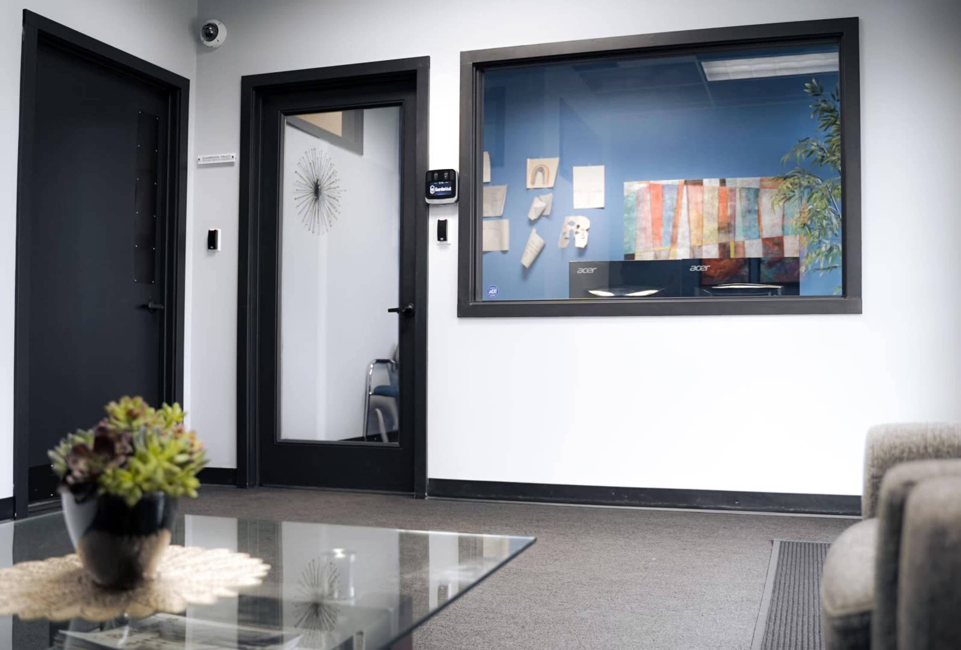 The image shows a modern office reception area featuring a glass wall with artwork, two black doors, and a coffee table with a small plant.