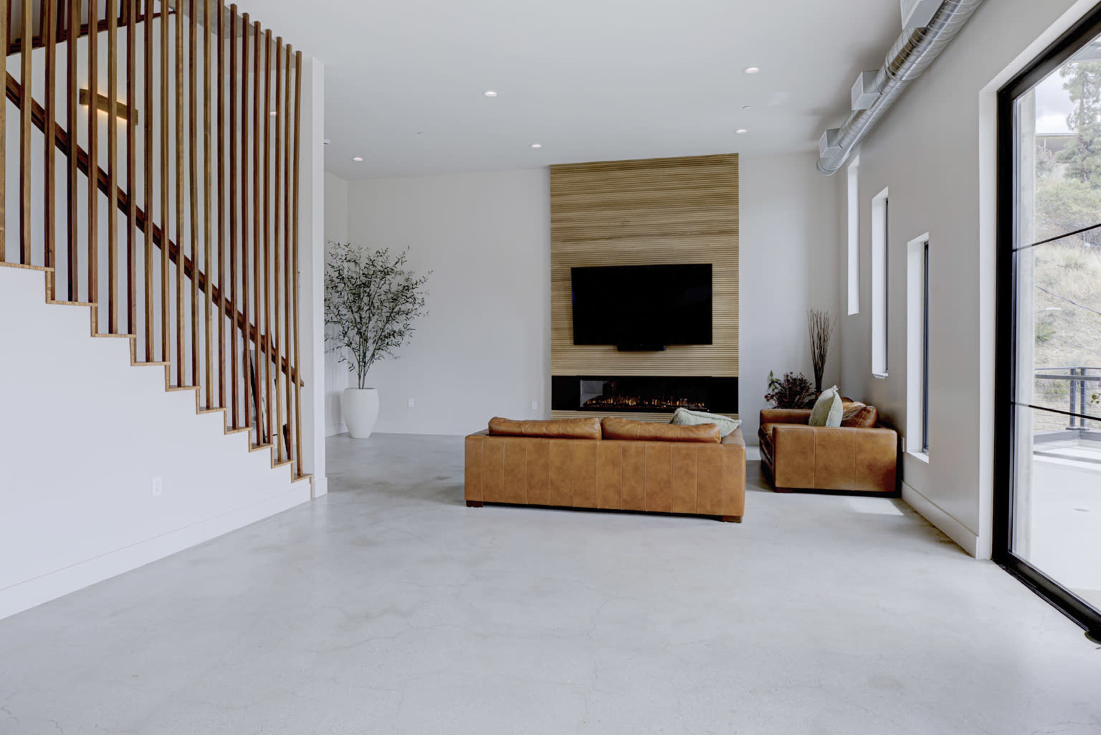 The image shows a spacious, modern living room with a leather sofa, a television mounted on a wooden accent wall, and a staircase with wooden railings.