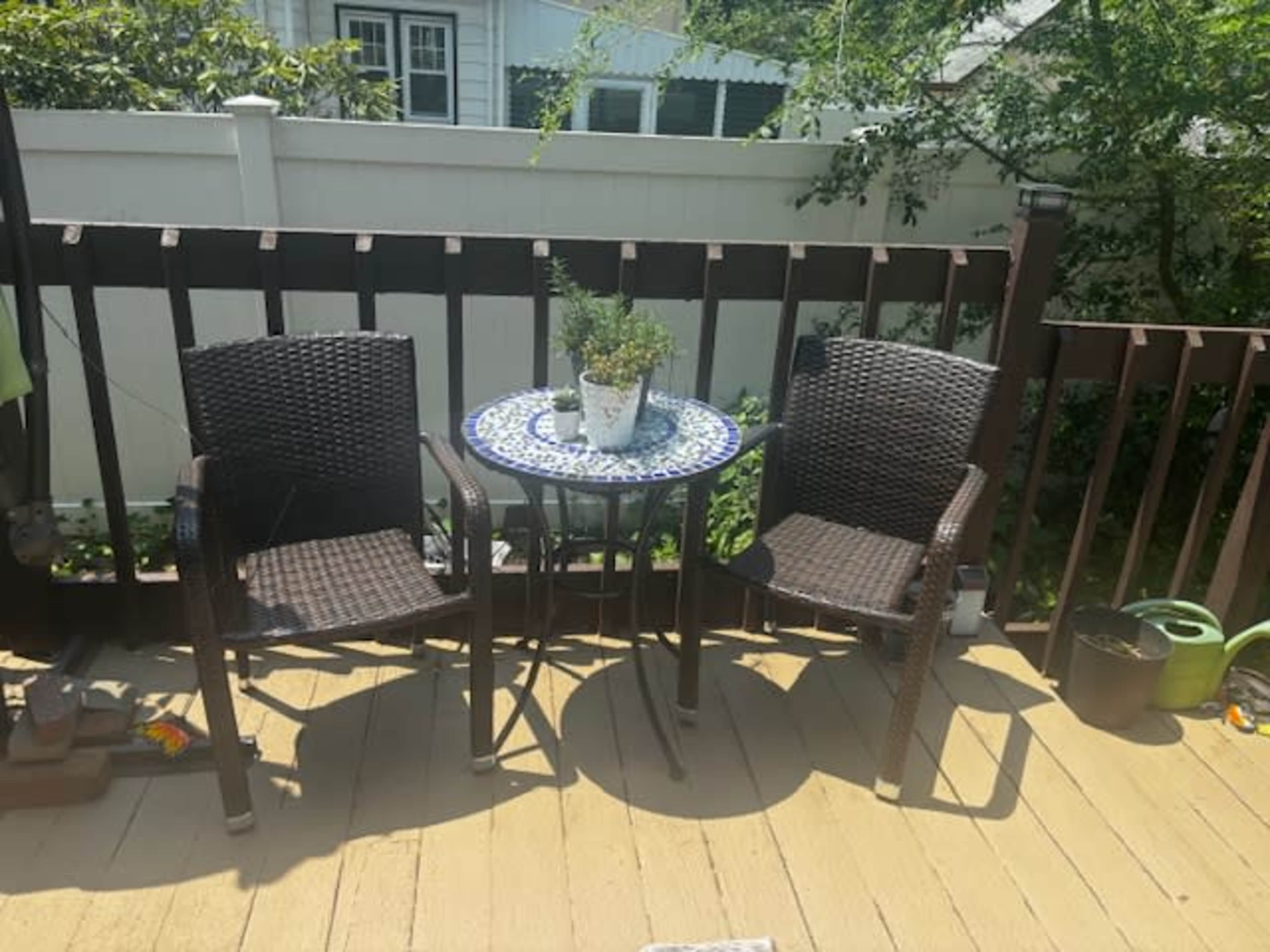Two wicker chairs are positioned beside a small round table with a potted plant on a wooden deck surrounded by greenery.
