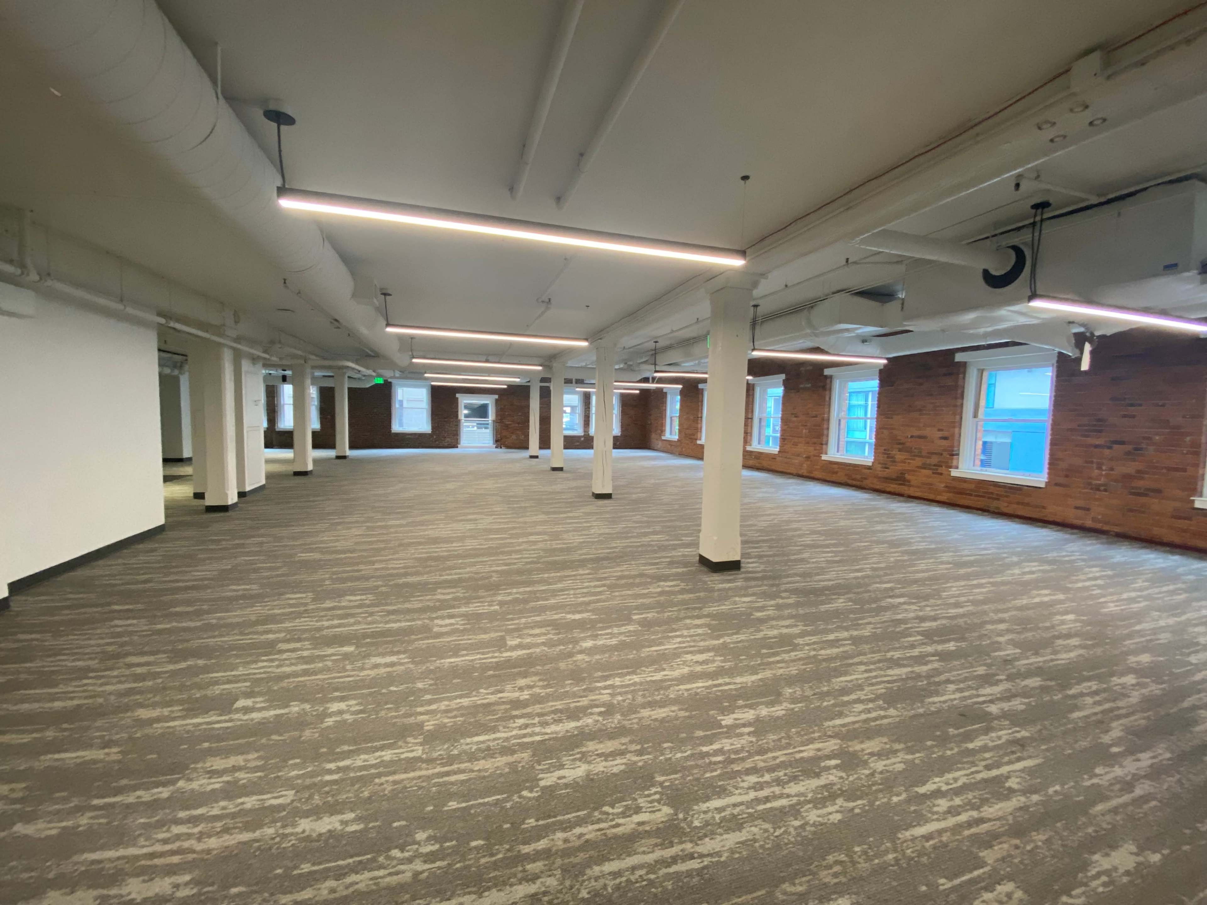 An empty office space features brick walls and large windows, illuminated by overhead fluorescent lights.