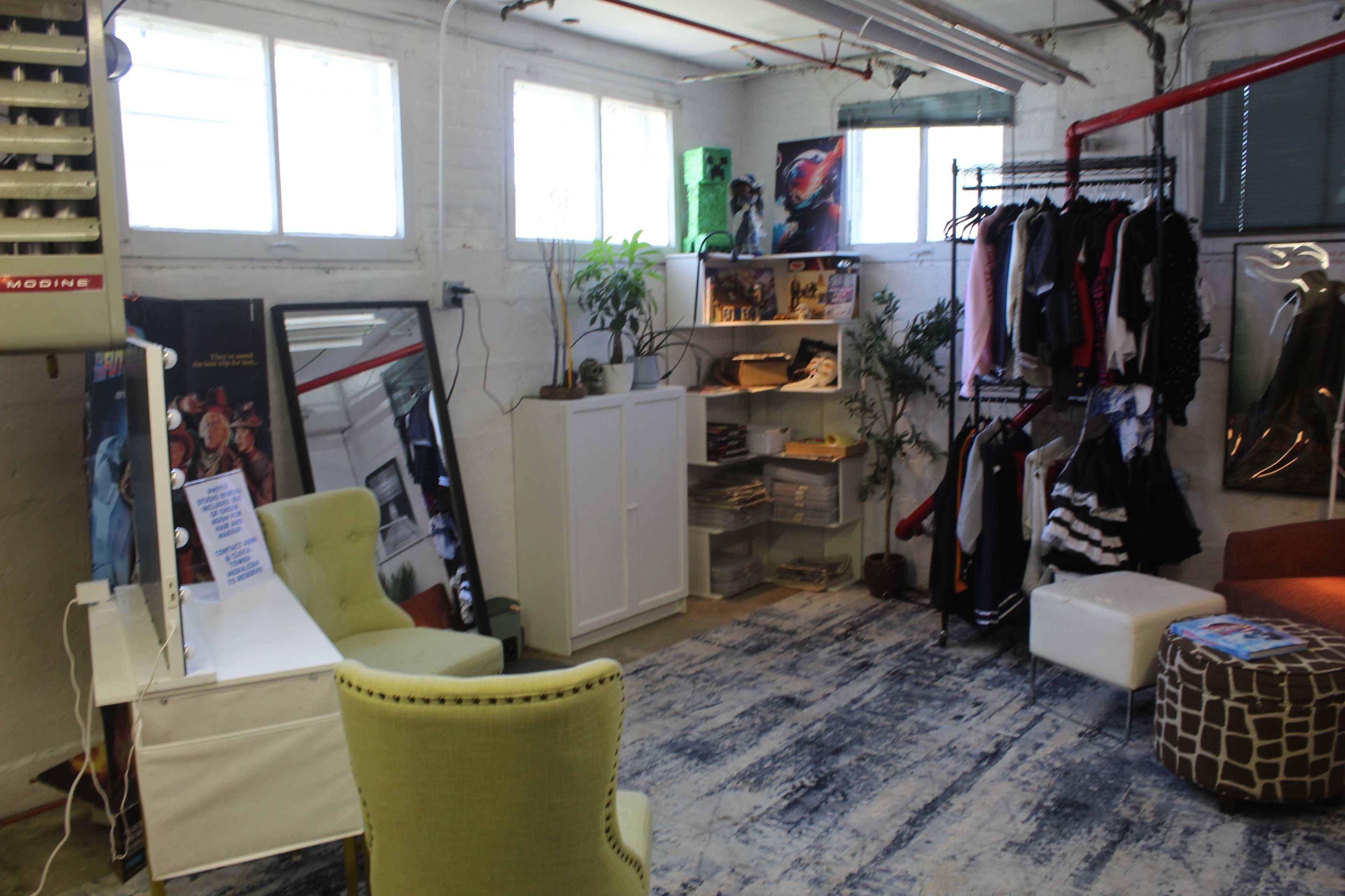 The image shows a small, well-organized basement space featuring clothing racks, green plants, a white cabinet, a TV setup, and an orange chair.