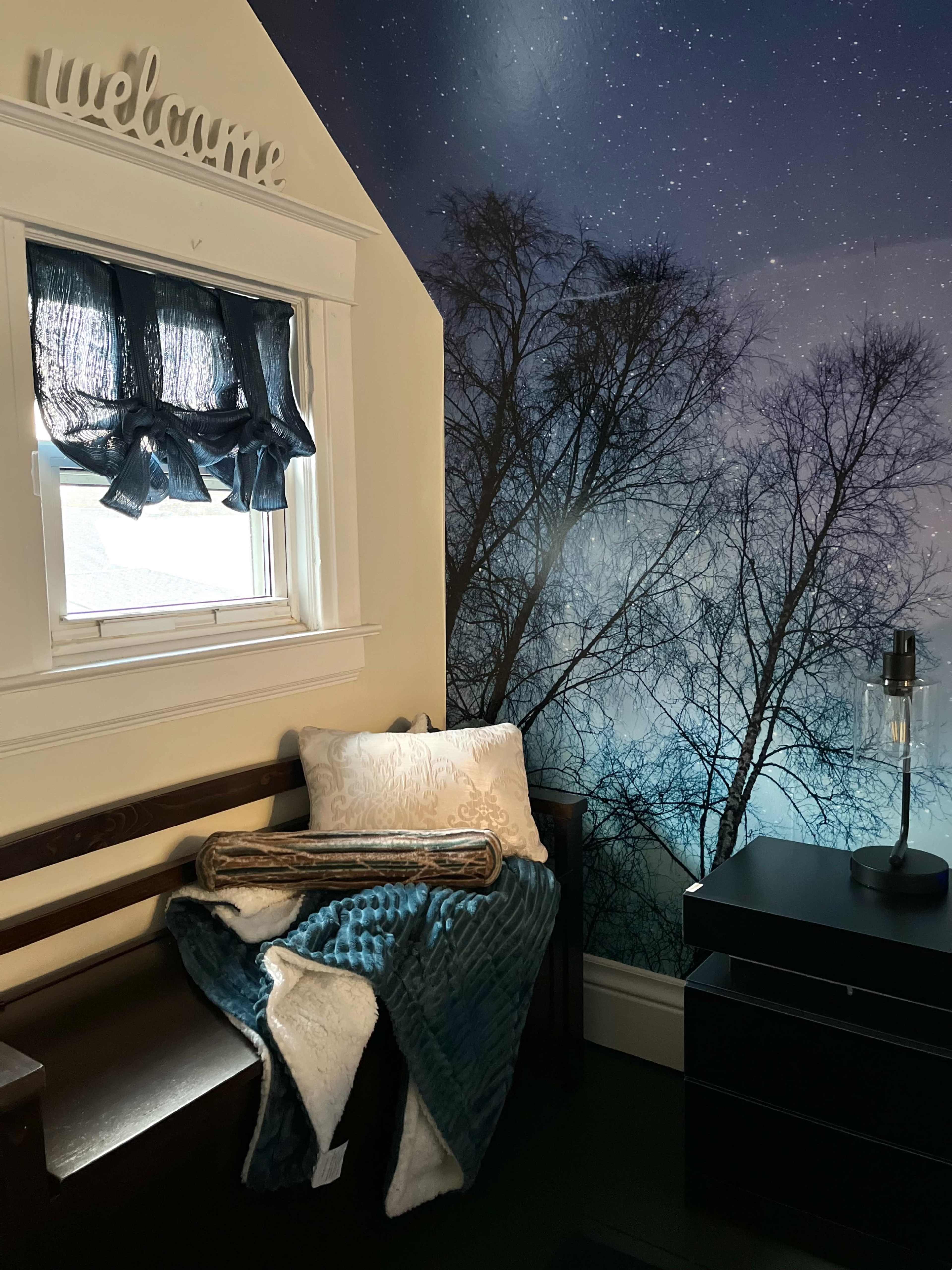 The image shows a cozy corner of a room with a window dressed in dark curtains, a bench with a decorative pillow and blanket, and a wall adorned with a nature-themed mural featuring a night sky and trees.