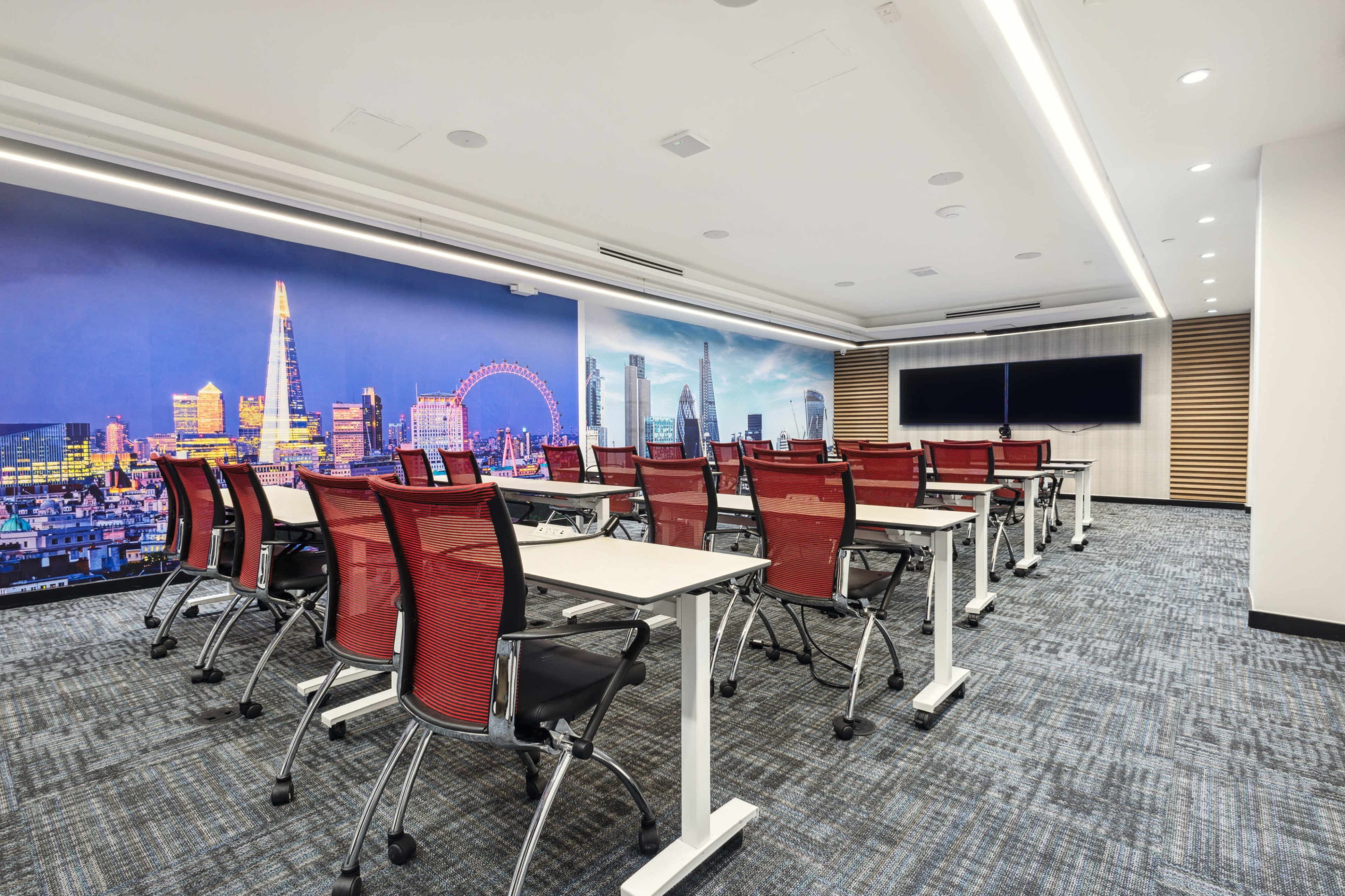 A modern conference room features rows of red office chairs and desks, with a large cityscape mural on the wall and a screen at the front.