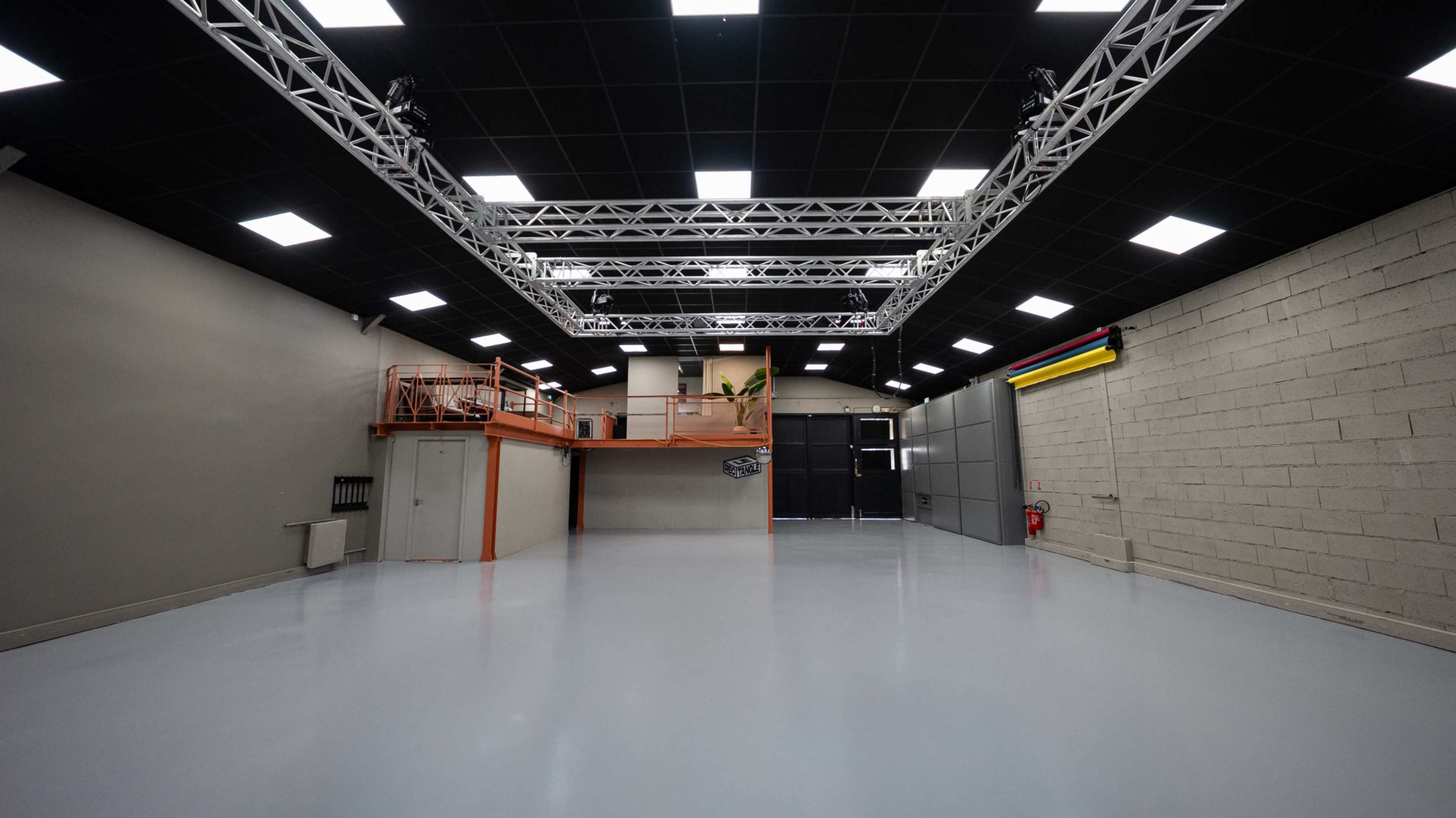 The image shows a spacious, empty studio or warehouse space with a gray floor, high ceilings, and a metal truss lighting setup.