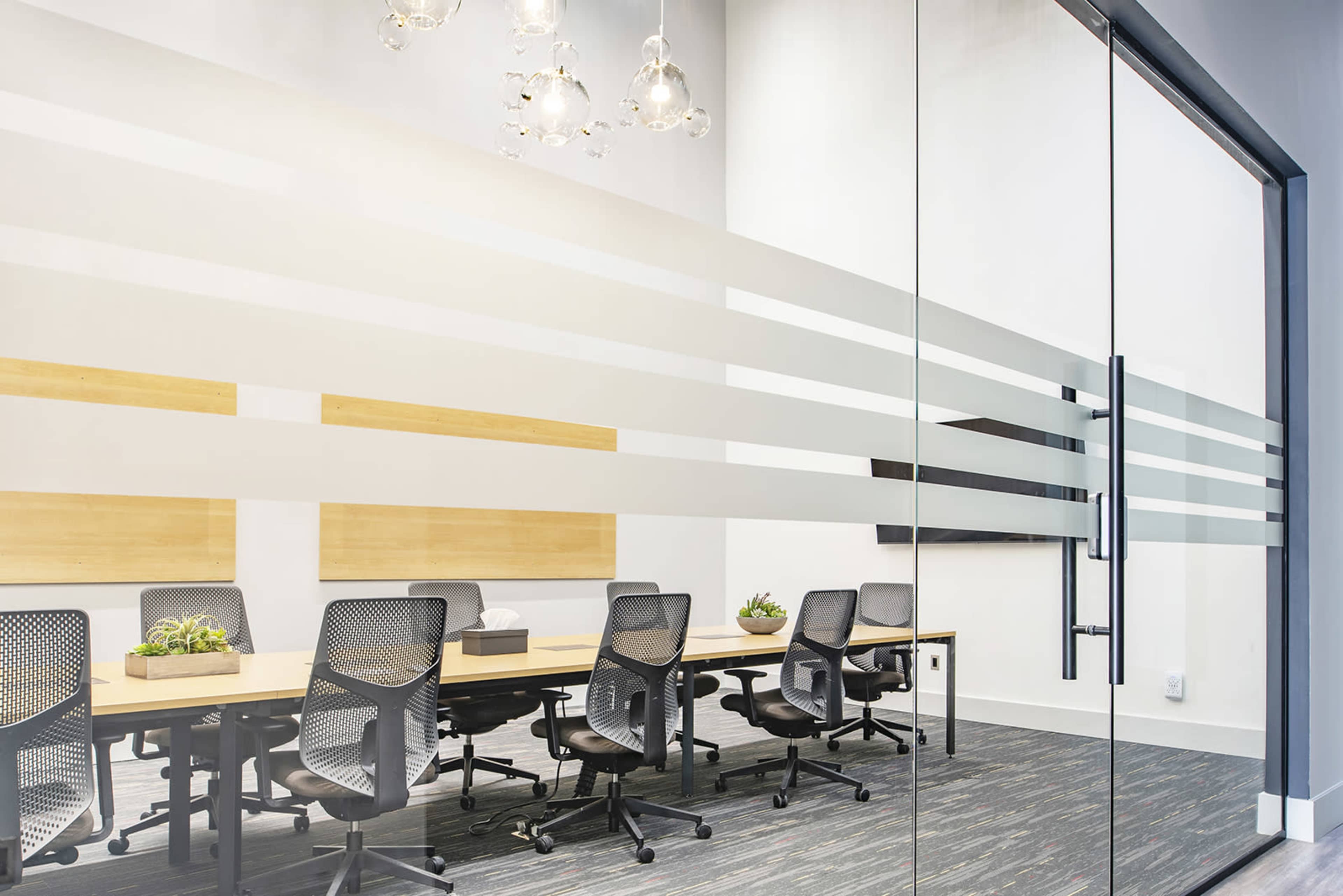 A modern conference room features a long table surrounded by several chairs, with large glass walls and decorative planters.