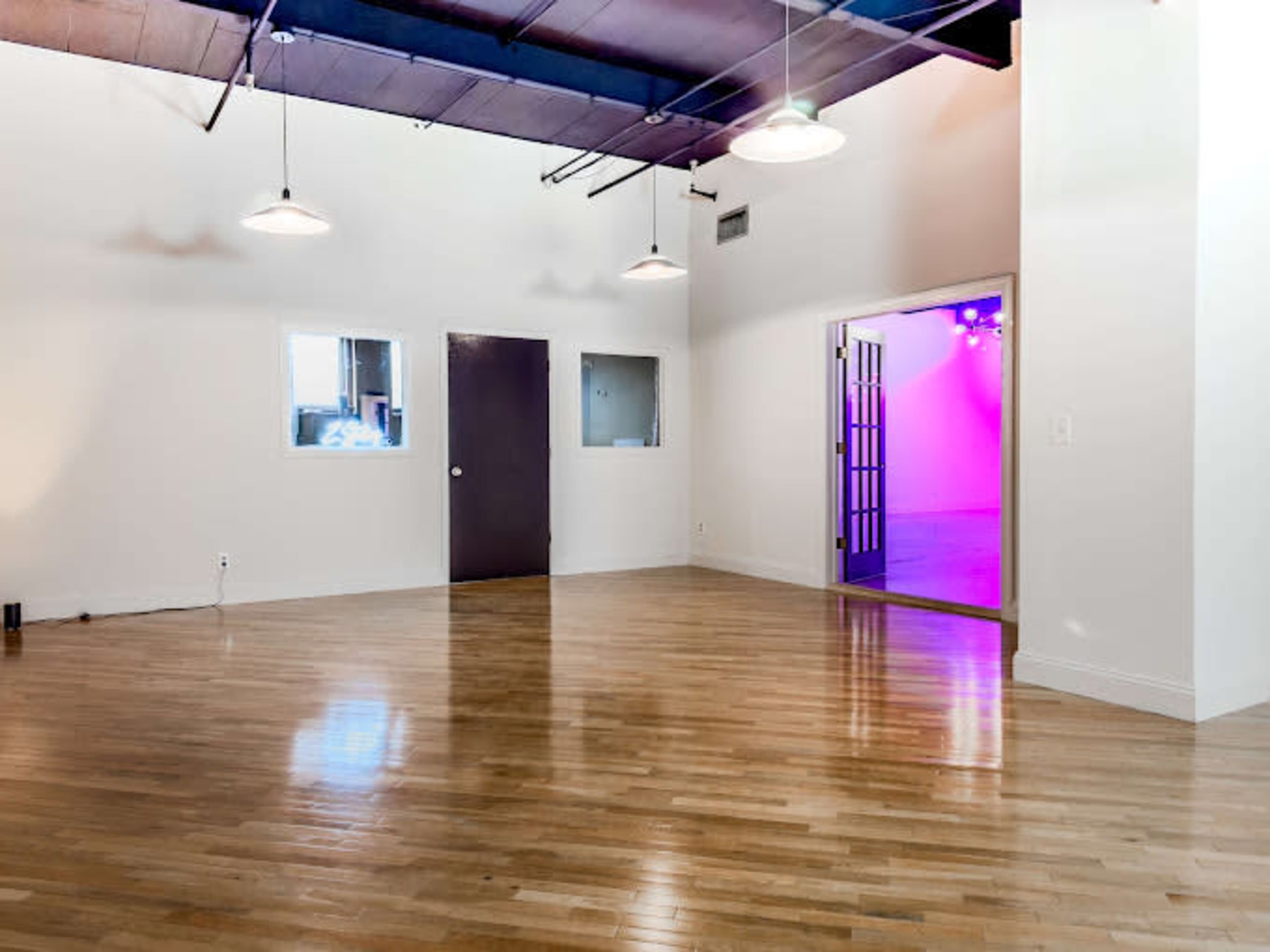 The image shows a spacious, well-lit room with hardwood floors, white walls, and a doorway leading to another room with colored lights.
