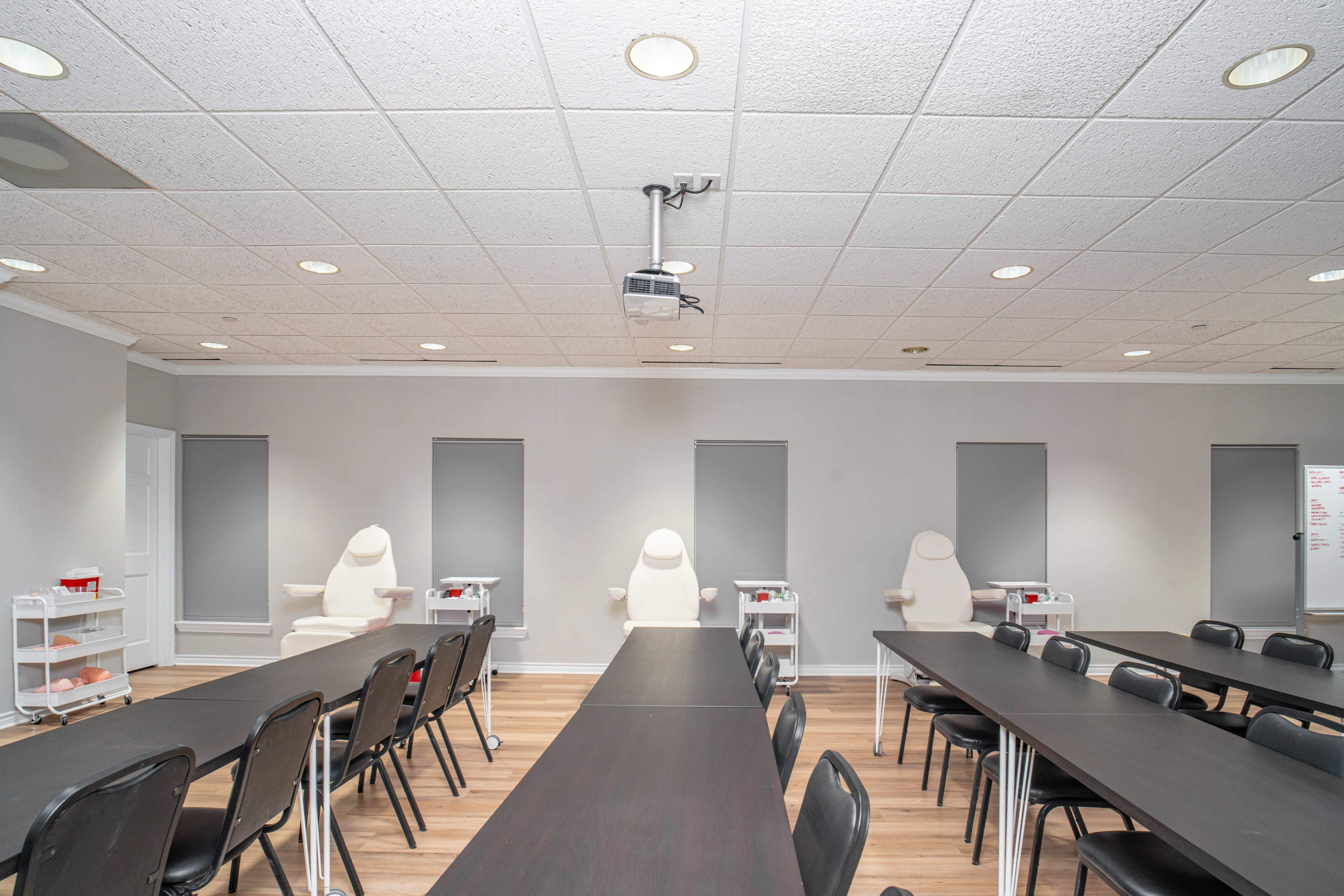 A training room with several long tables, chairs, and three examination chairs at the front, equipped with a projector and closed window shades.
