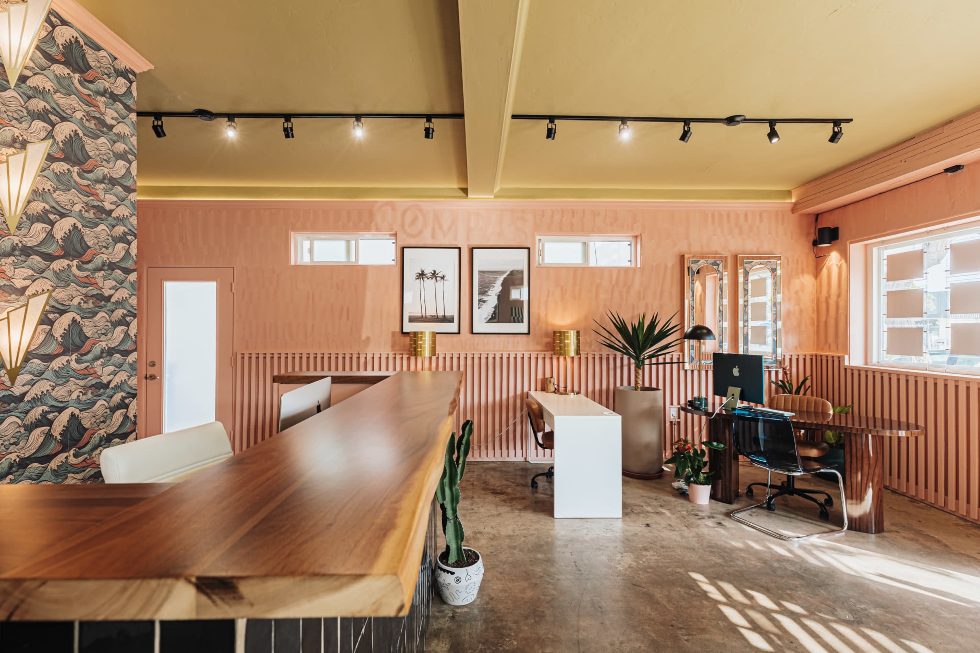 The image shows a modern office reception area with a wooden desk, a minimalist white workspace, and decorative plants against pink walls adorned with patterned wallpaper.