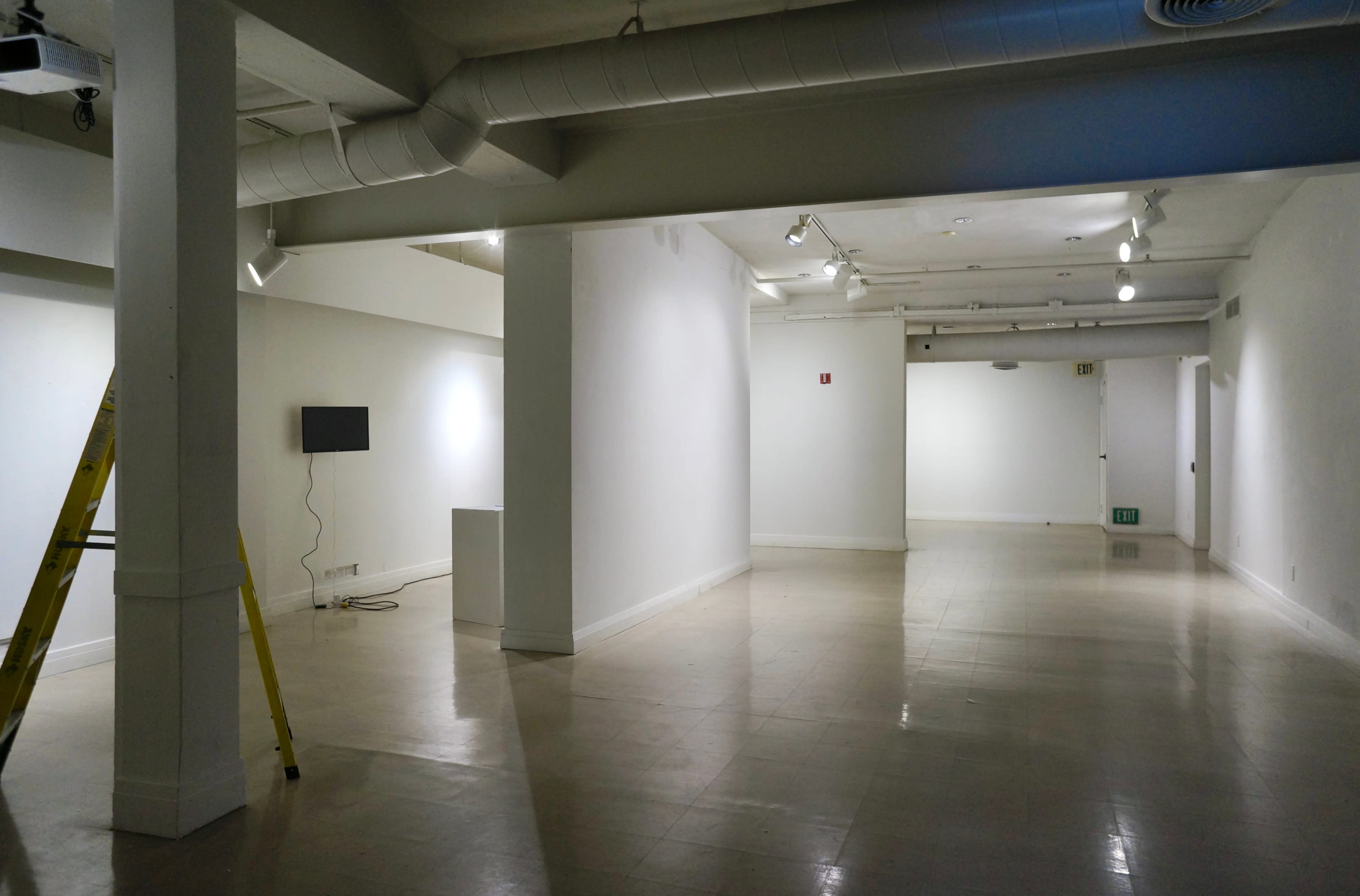 A spacious, well-lit art gallery with white walls, a television on one wall, and a ladder in the foreground.