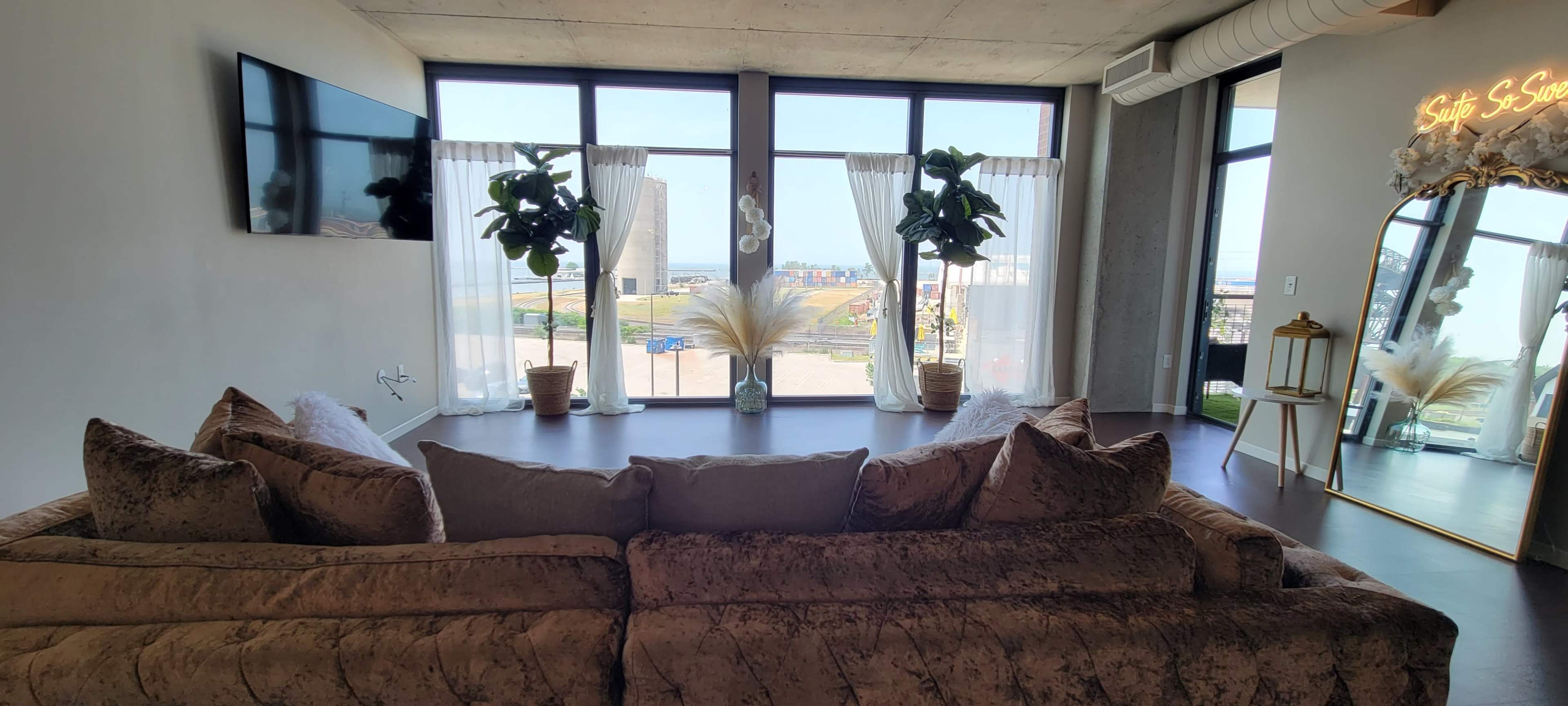 A modern living room with a large window revealing a view of an outdoor area, featuring a plush sofa, decorative plants, and sheer curtains.