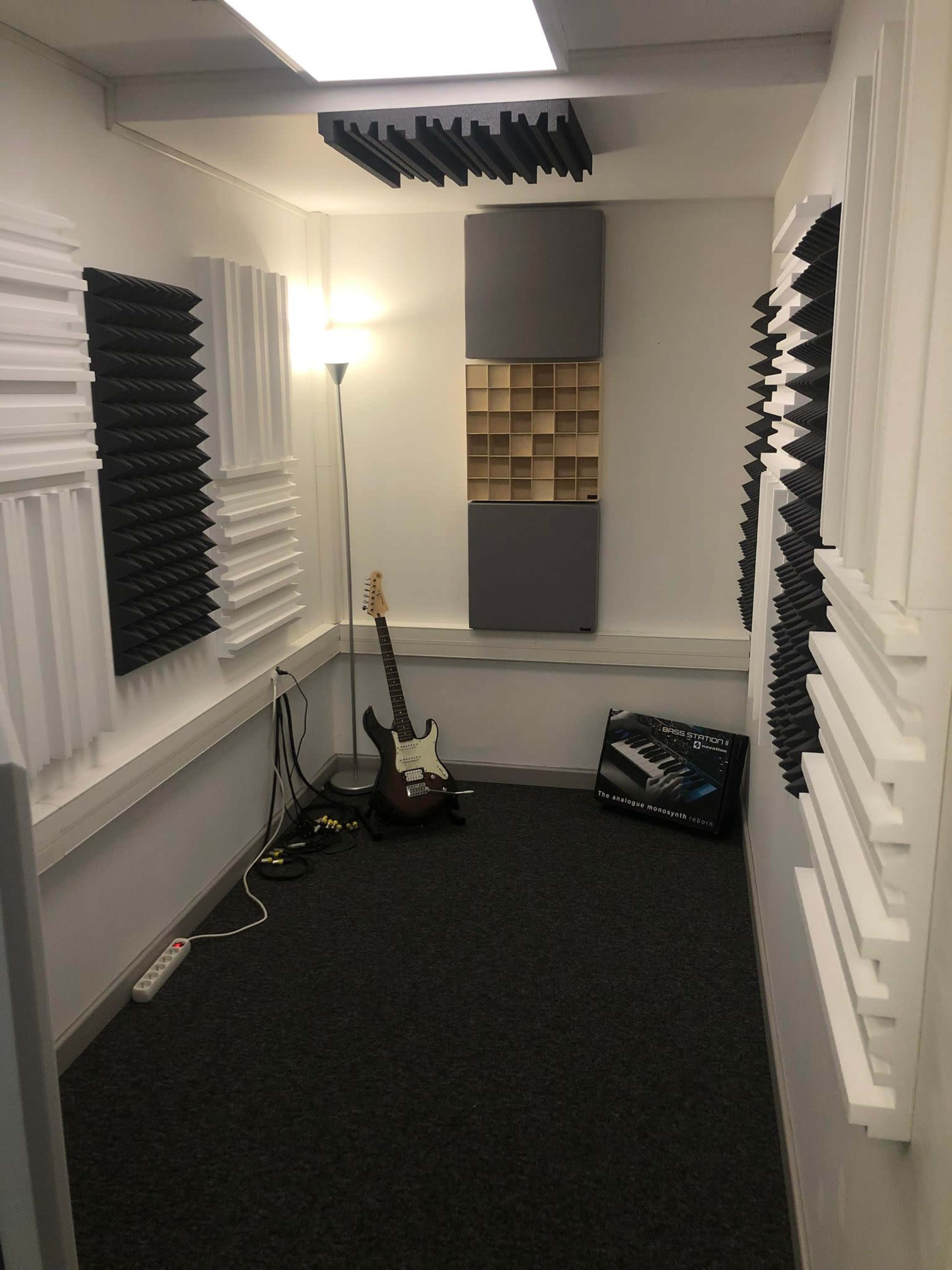 A small recording studio with soundproofing panels on the walls, a lamp, an electric guitar, and a keyboard case on the floor.
