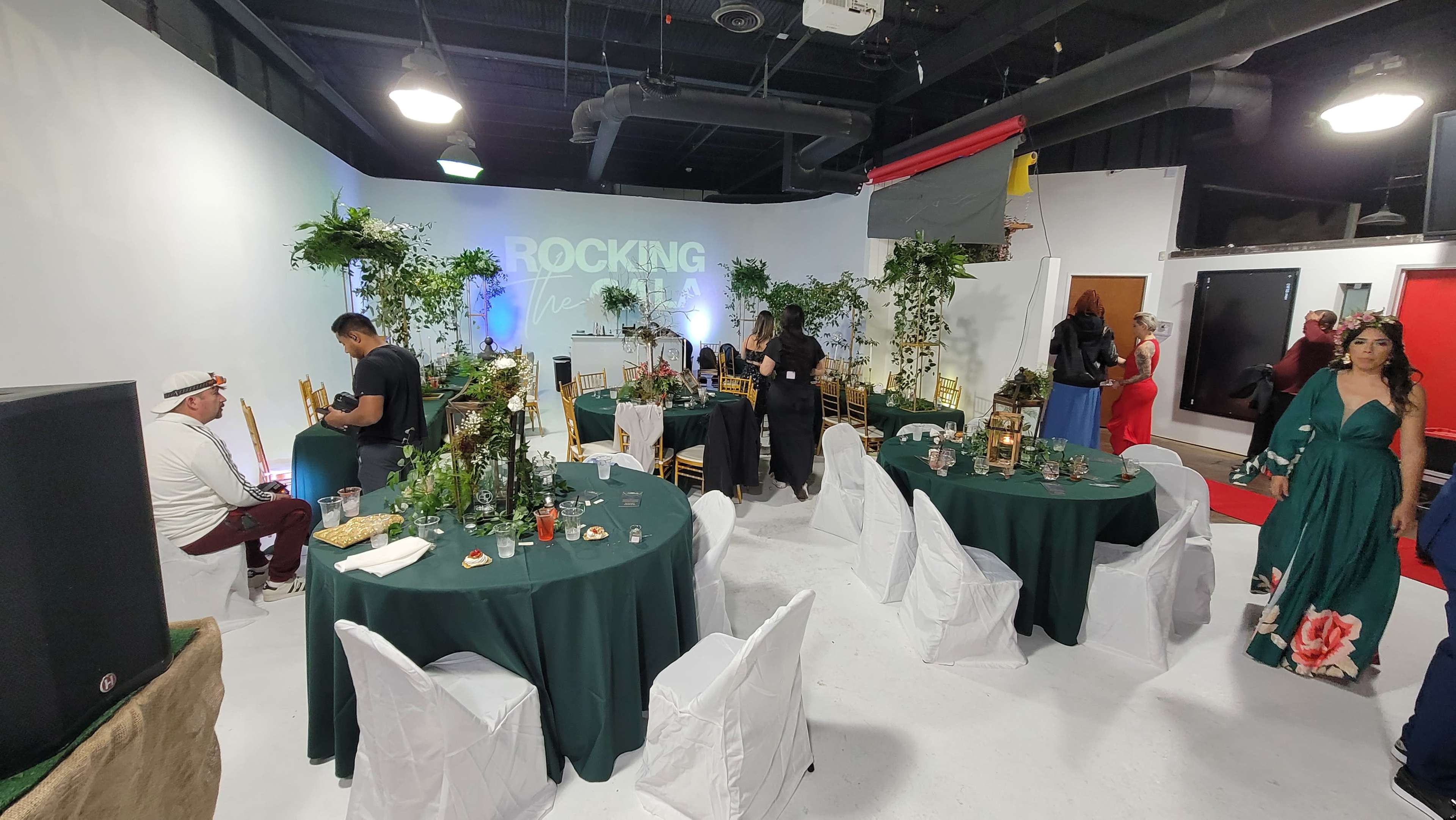 A spacious event venue features round tables set with green tablecloths, surrounded by guests mingling among decorative plants and a backdrop that reads "ROCKING THE V".