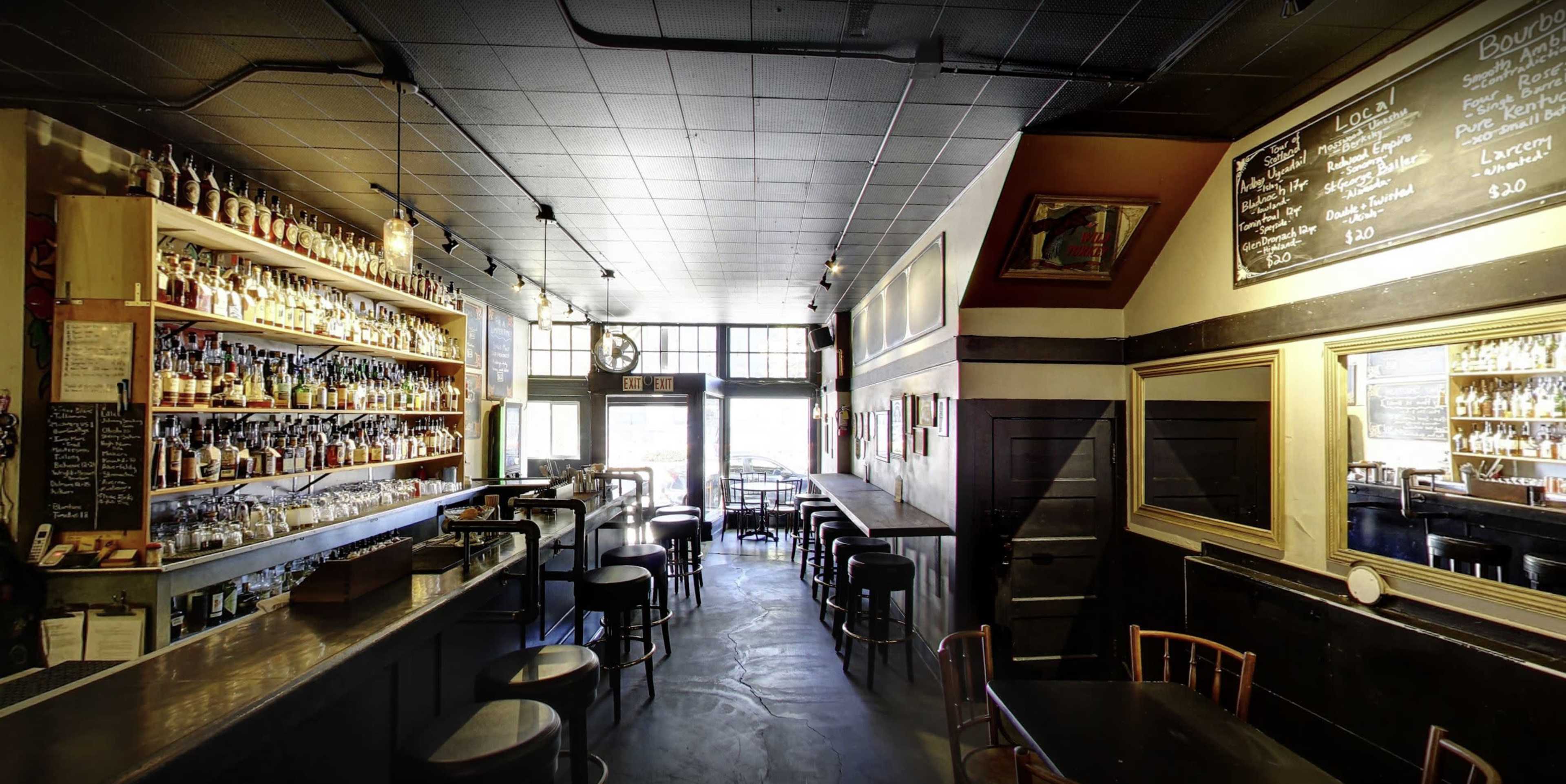 A dimly lit bar with a long wooden counter, shelves lined with various bottles, and empty tables and chairs facing large windows.