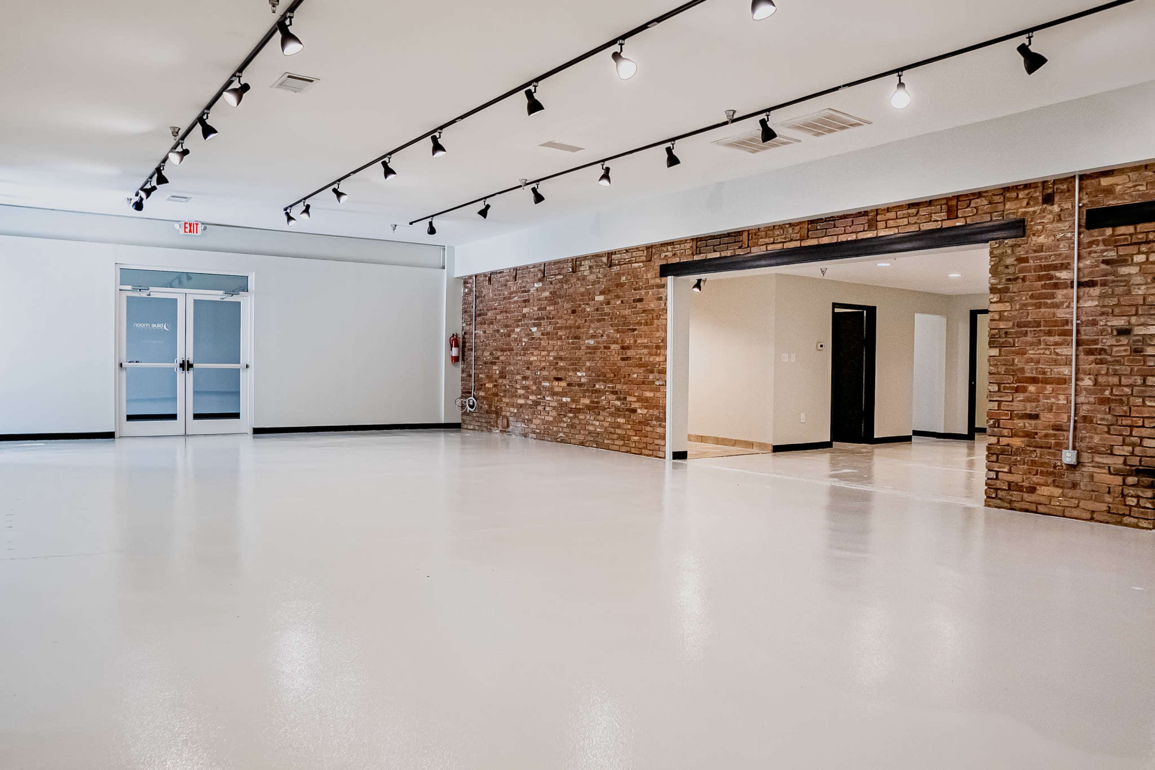 An empty room with polished white floors, exposed brick walls on one side, and track lighting along the ceiling.