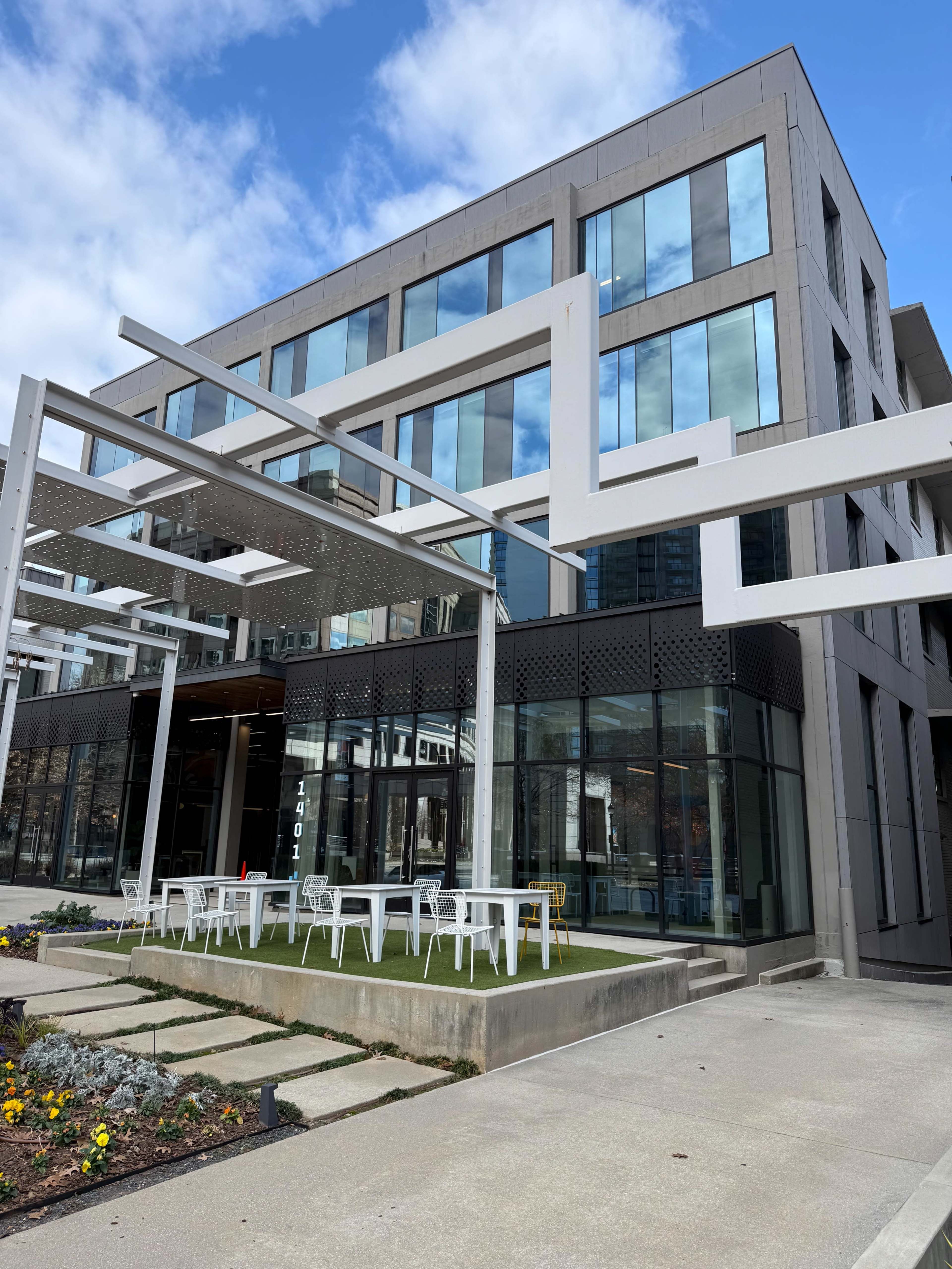 The image shows a modern, multi-story building with large glass windows and an outdoor seating area featuring white tables and chairs under a geometric awning.