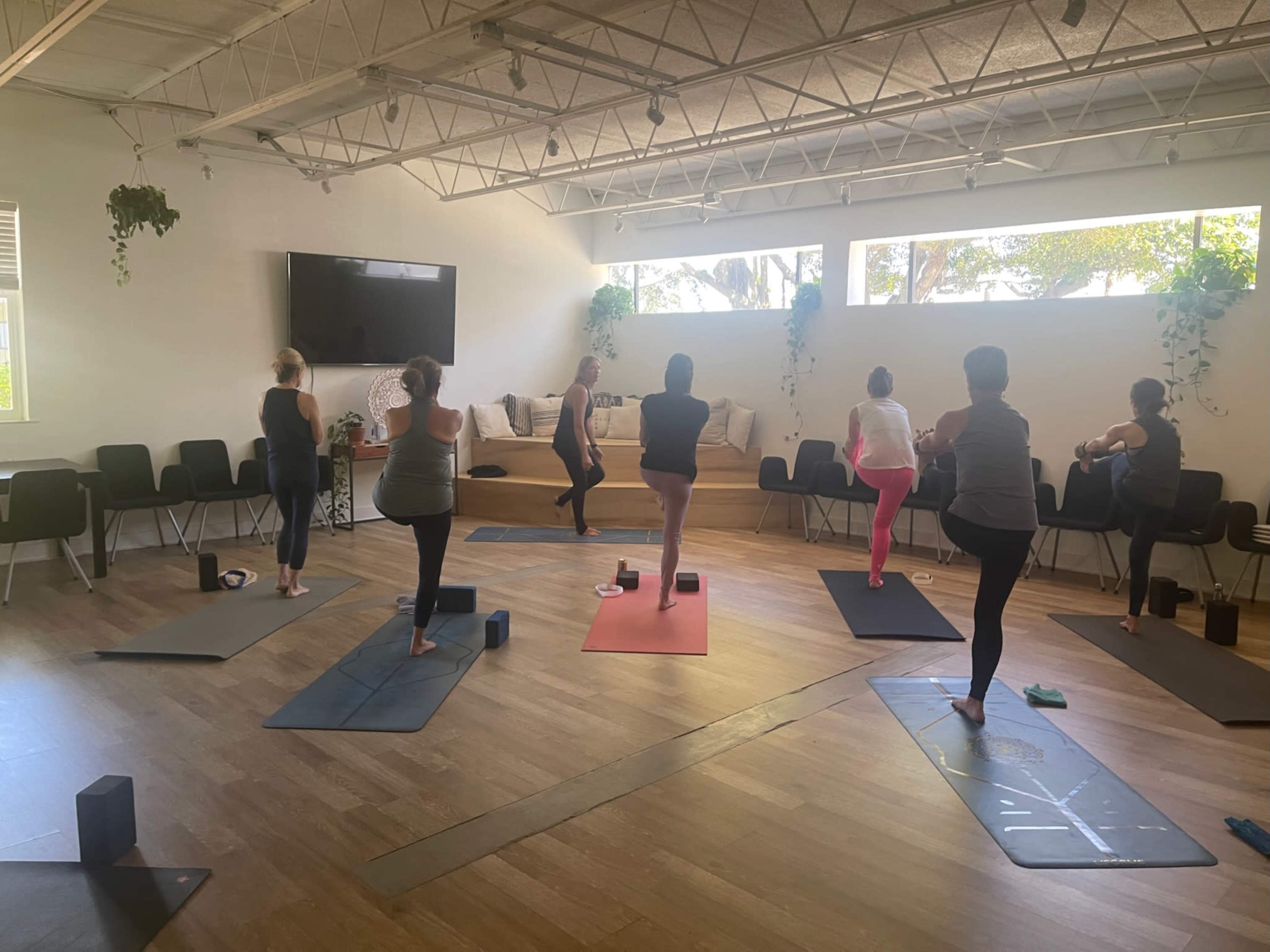 A group of people practicing yoga in a well-lit studio with wooden floors and mats arranged on the ground.