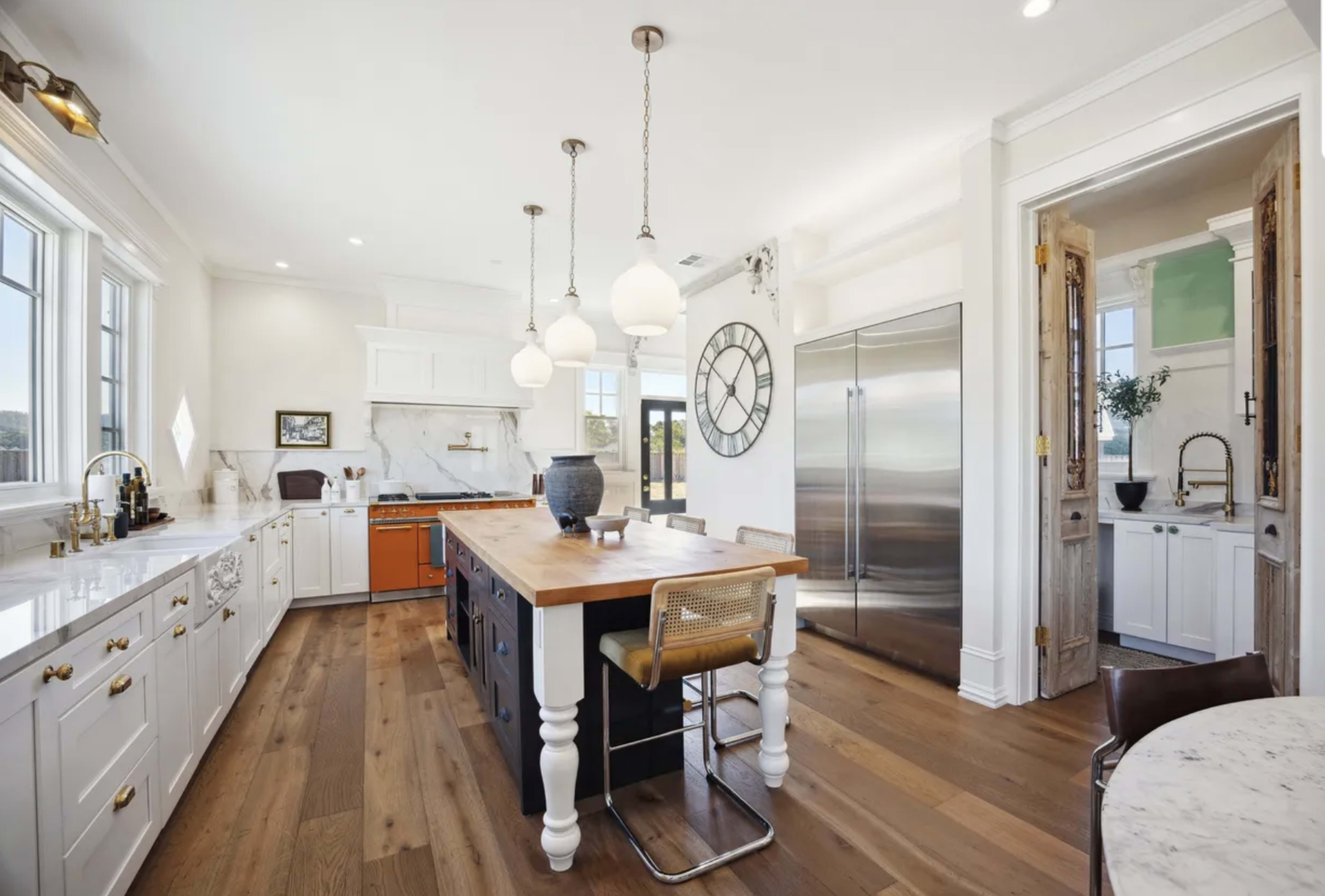 The kitchen features a central island with a wooden top, white cabinets, stainless steel appliances, and large windows allowing natural light to illuminate the space.