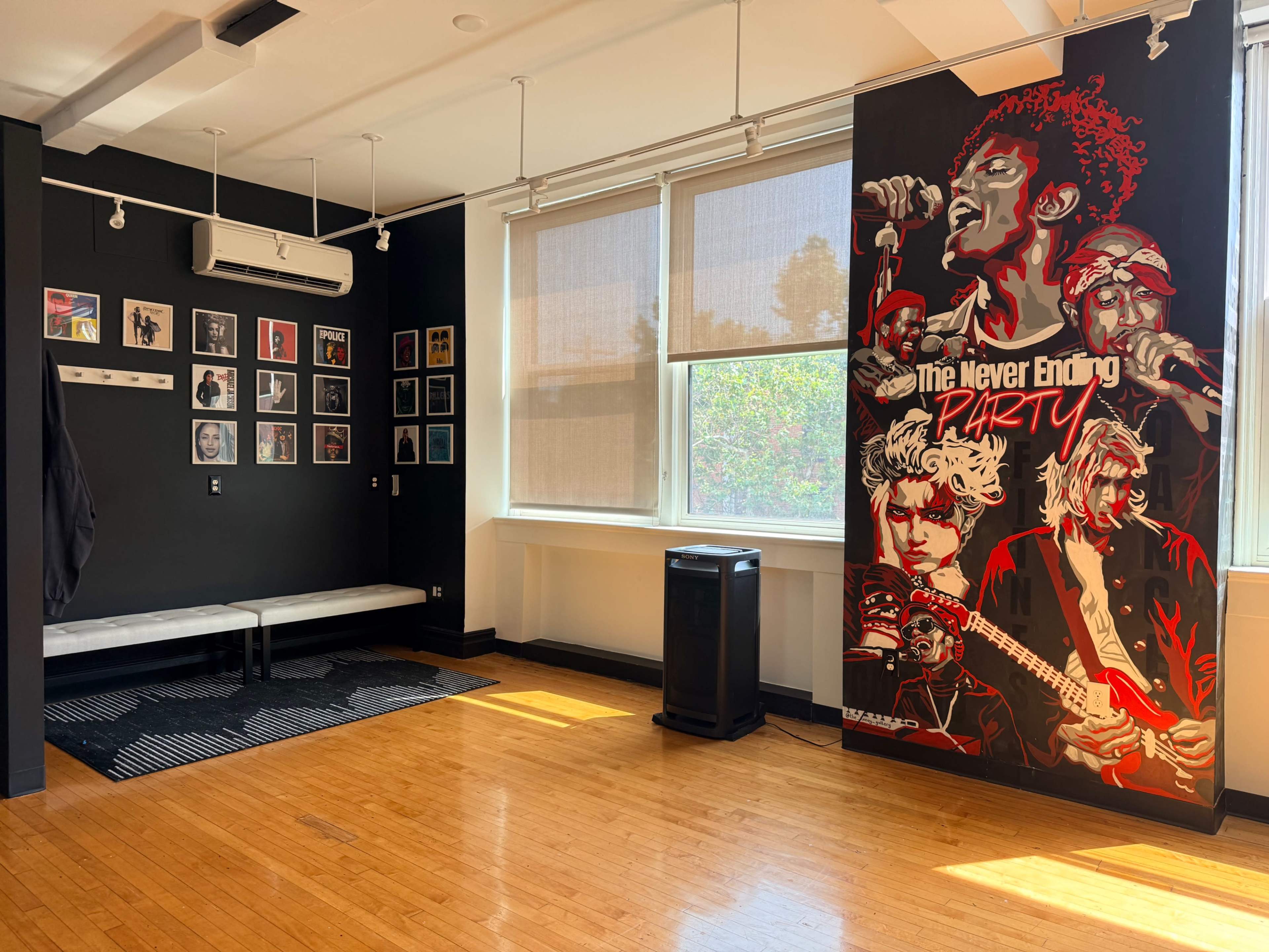 The room features a black wall adorned with framed art and a large, colorful mural of musicians, alongside a wooden floor and windows with shades.