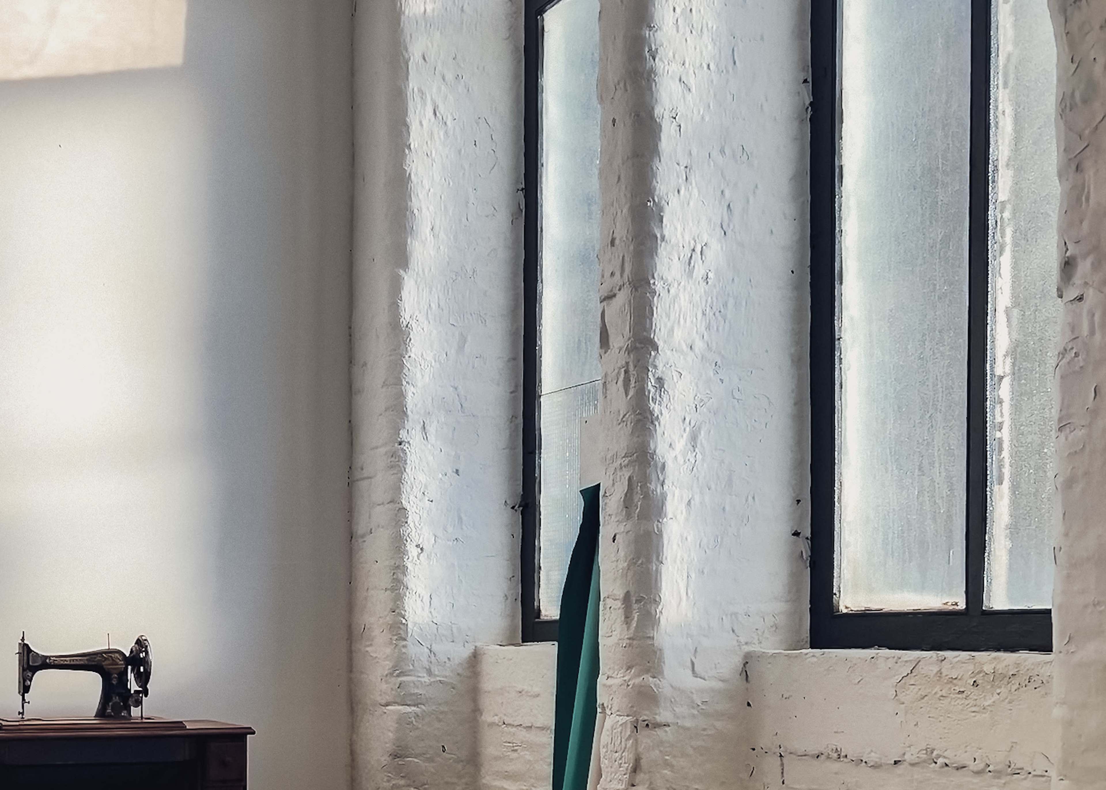 The image shows a room with large windows allowing light to enter, a vintage sewing machine on a wooden table, and a piece of fabric draped against a wall.