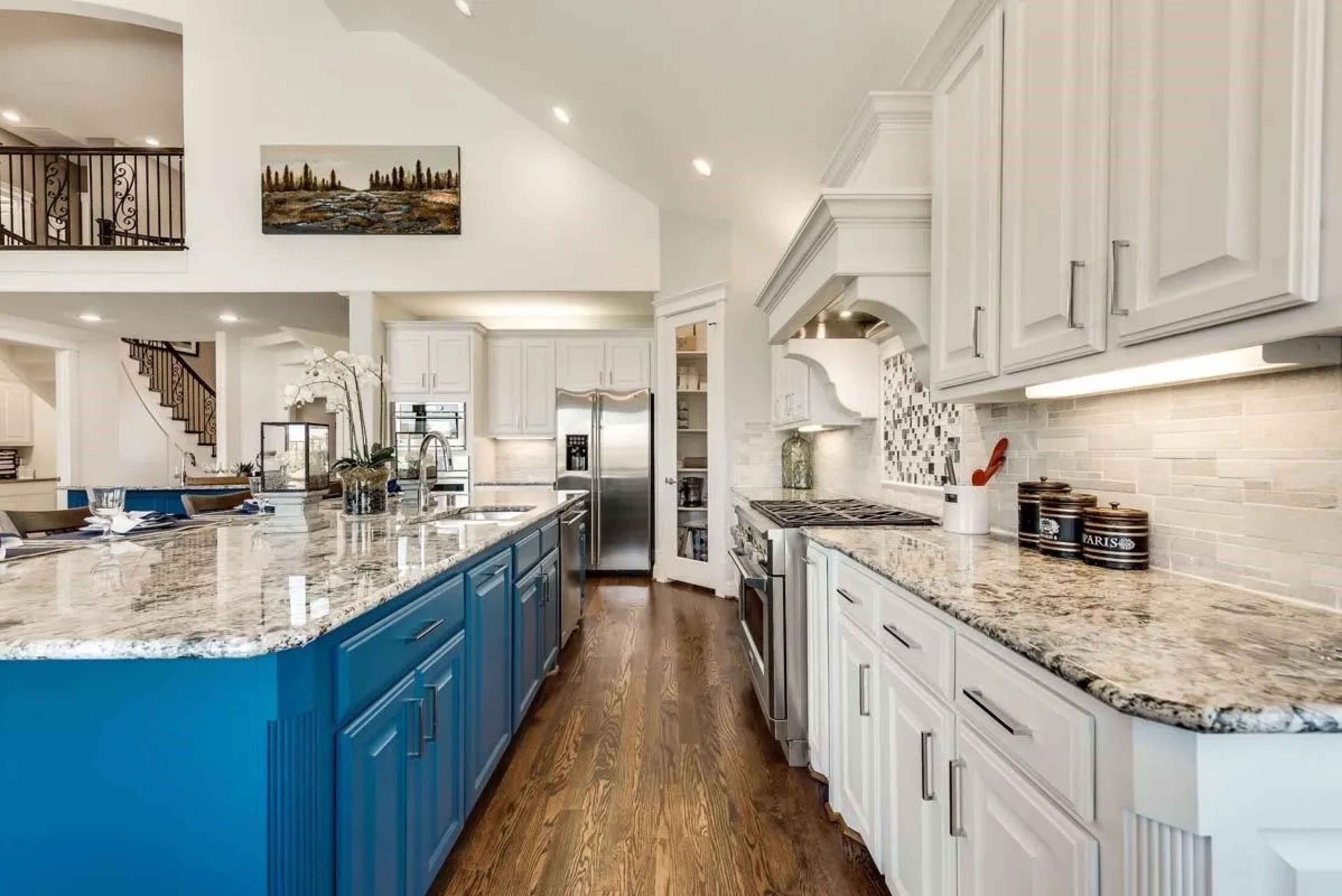 The image shows a modern kitchen with white cabinetry and blue accent cabinets, featuring a marble countertop and stainless steel appliances.