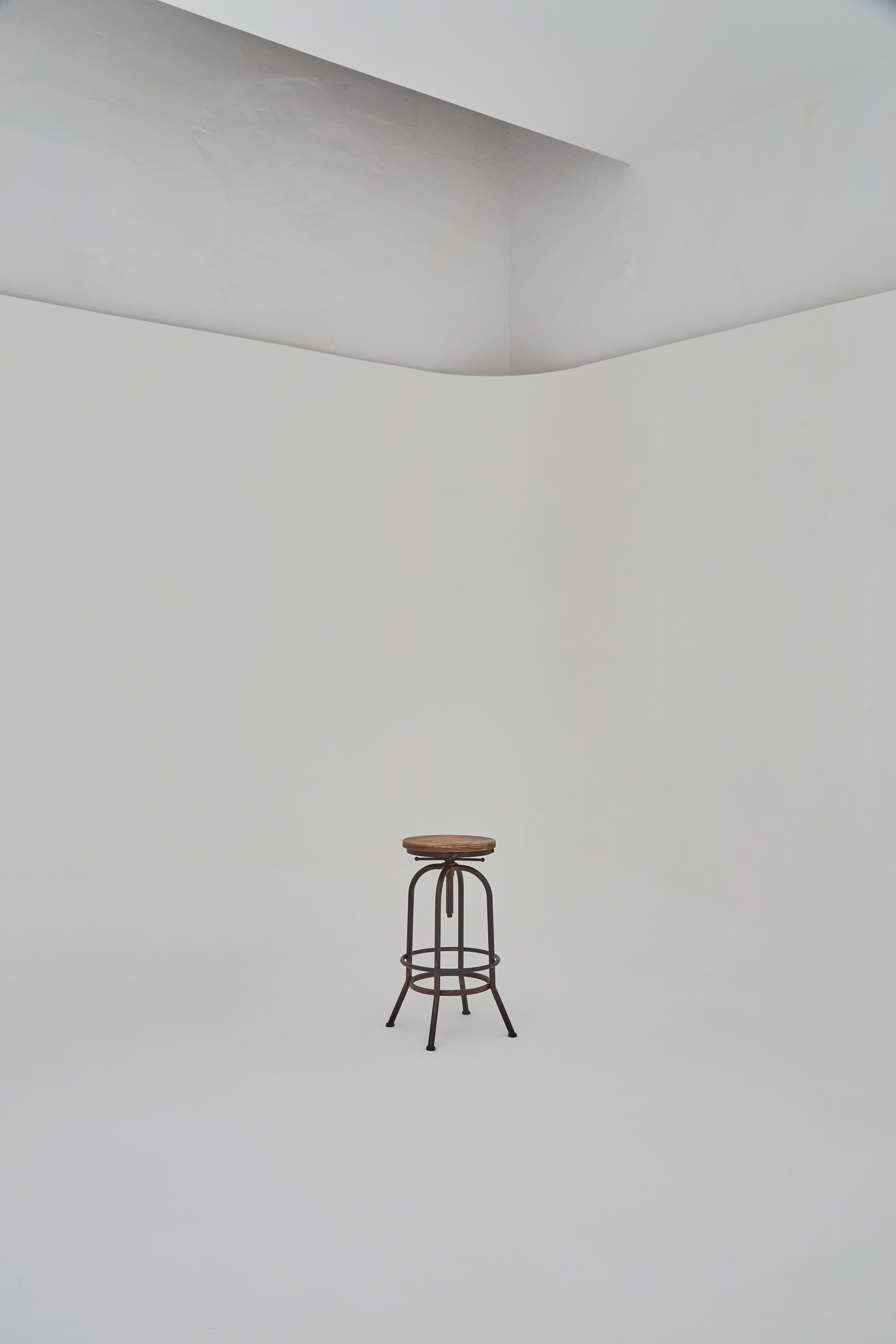 A metal and wood bar stool stands alone on a white floor in an empty room with white walls.