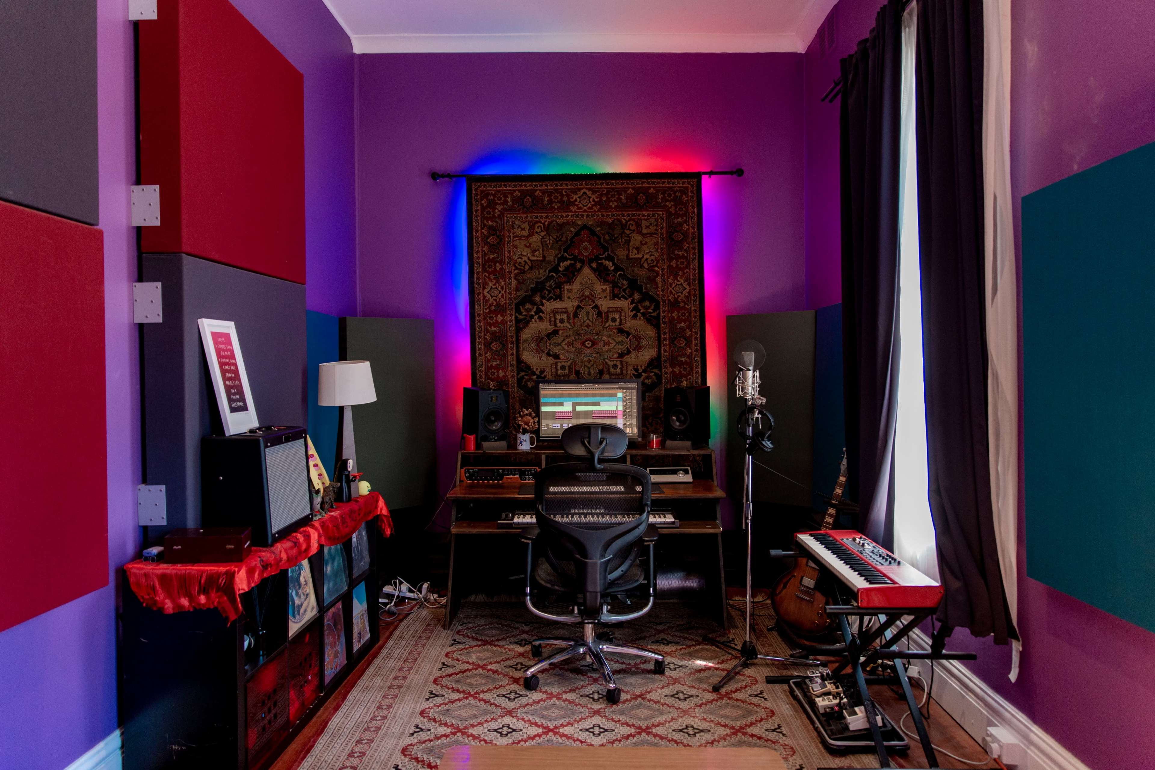 A music production studio features a desk with a computer, a microphone, and colorful soundproofing panels on the walls.